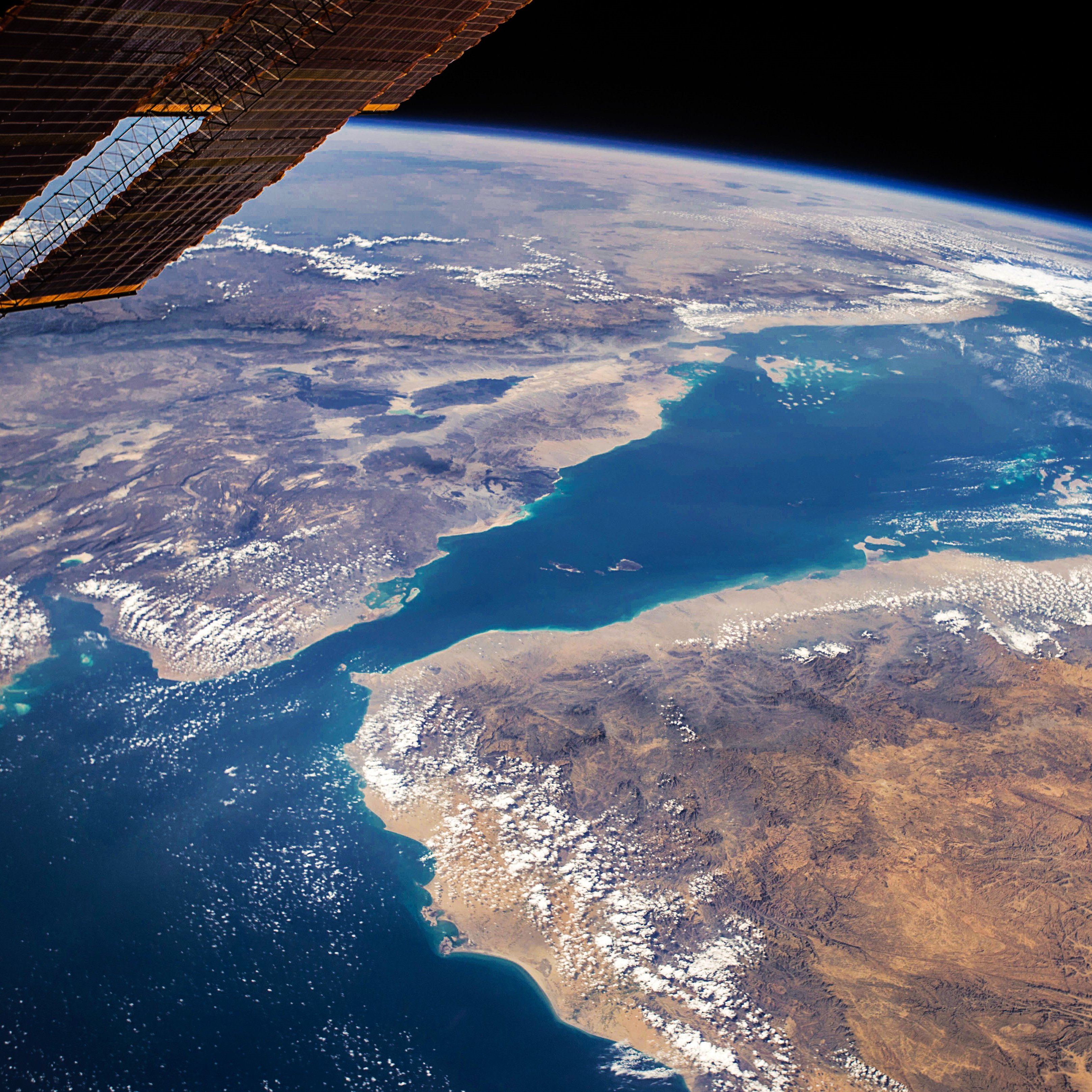 Cette image, prise depuis l'espace, montre une vue spectaculaire de la Terre. On y voit des paysages variés, avec une mer d'un bleu profond bordée de terres aux couleurs chaudes, tantôt sableuses, tantôt montagneuses. Il y a des nuages éparpillés qui ajoutent du relief à la scène. Les contours des côtes et des îles se dessinent nettement. En haut de l'image, on peut apercevoir la noirceur de l'espace, ce qui crée un beau contraste avec les teintes vives de la planète. Cette vue donne une impression de sérénité et d'immensité, rappelant la beauté fragile de notre environnement.