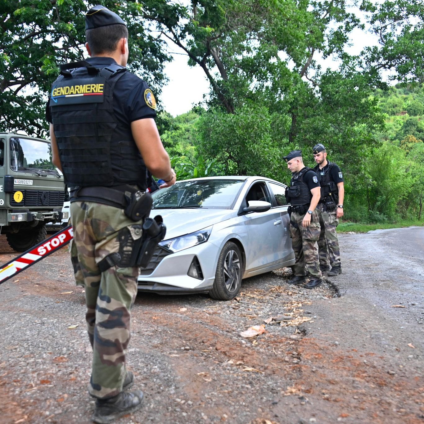 L'image montre un contrôle routier en plein air. Au premier plan, un gendarme en uniforme, portant un gilet par balles noir avec l'inscription "Gendarmerie", observe une voiture argentée qui est arrêtée sur le côté de la route. La voiture a un panneau "STOP" visible. Deux autres gendarmes, habillés de manière similaire, se tiennent près de la voiture, dialoguant probablement avec le conducteur. En arrière-plan, on aperçoit une camionnette militaire de la gendarmerie et des arbres verdoyants qui donnent une ambiance naturelle à la scène. Le sol est en terre, avec beaucoup de verdure autour, ce qui indique que le lieu est rural. L'atmosphère semble sérieuse, reflet du contrôle de sécurité en cours.