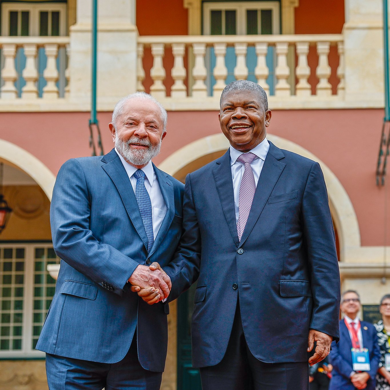 L'image montre deux hommes se serrant la main, symbolisant un moment de convivialité et de coopération. En arrière-plan, on peut apercevoir une architecture colorée avec des colonnes et des balcons. Des personnes sont rassemblées à l'arrière, témoignant d'un événement officiel ou d'une rencontre importante. Les vêtements des hommes sont élégants : l'un porte un costume gris et l'autre un costume noir avec une cravate claire. L'atmosphère semble chaleureuse et dynamique, reflétant un échange amical.