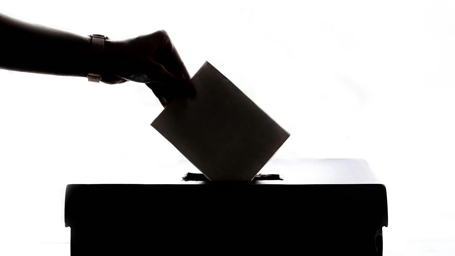 L'image représente une scène de vote. On peut voir une main tendue vers une urne, prête à y insérer un bulletin de vote. La silhouette de la main et de l'urne est visible contre un fond lumineux, créant un contraste fort. L'urne est de forme rectangulaire et se trouve sur une surface plane. Cette image évoque le processus démocratique et l'importance de l'engagement civique.
