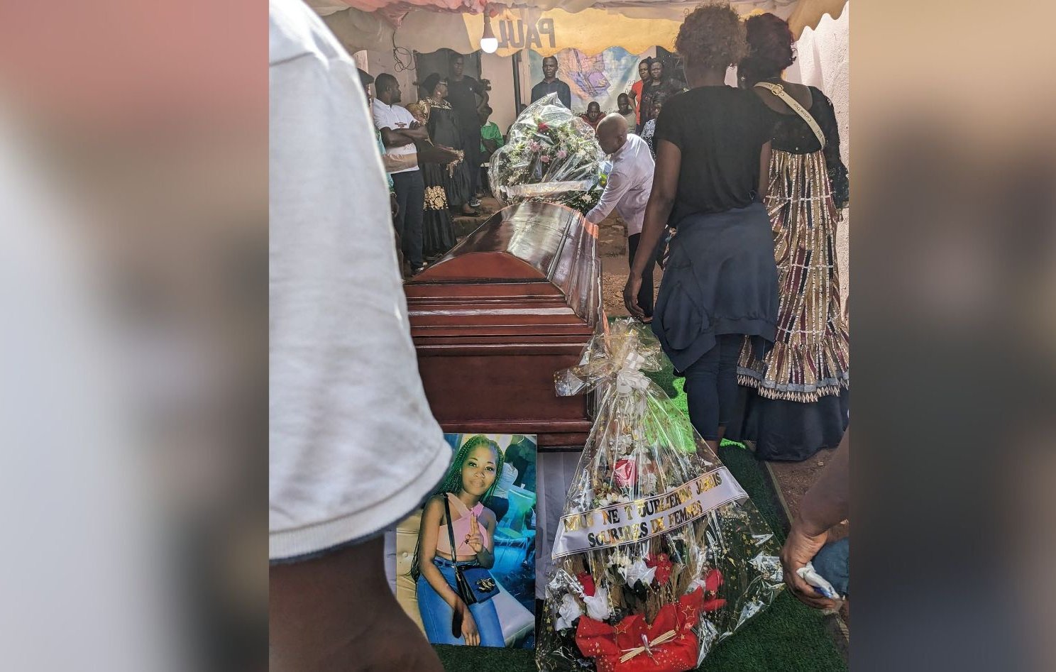 L'image montre un moment poignant d'un enterrement. Au centre, un cercueil en bois brun, sur lequel sont déposés des bouquets de fleurs. À côté du cercueil, une grande photo d'une femme est accrochée, entourée de décorations florales et de bandes de tissu. Des personnes, visiblement en deuil, se rassemblent autour, certaines portant des vêtements sombres. L'ambiance est sombre mais respectueuse, marquée par la tristesse et l'hommage à la personne décédée. L'arrière-plan est flou, mais on peut deviner une tente qui abrite cette cérémonie. L'ensemble évoque des sentiments de perte et de commémoration.