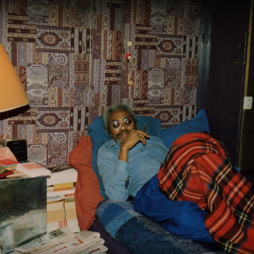 L'image montre un homme allongé sur un lit, recouvert d'une couverture à carreaux rouges et noirs. Il est vêtu d'une chemise bleue et porte des lunettes. L'homme semble pensif, avec un léger sourire. En arrière-plan, on distingue un mur tapissé d'un motif coloré et complexe. À côté du lit, une lampe de table en métal émet une lumière douce. Il y a également des piles de livres et de journaux sur une table à proximité, ce qui donne une impression de confort et d'intimité dans la pièce.