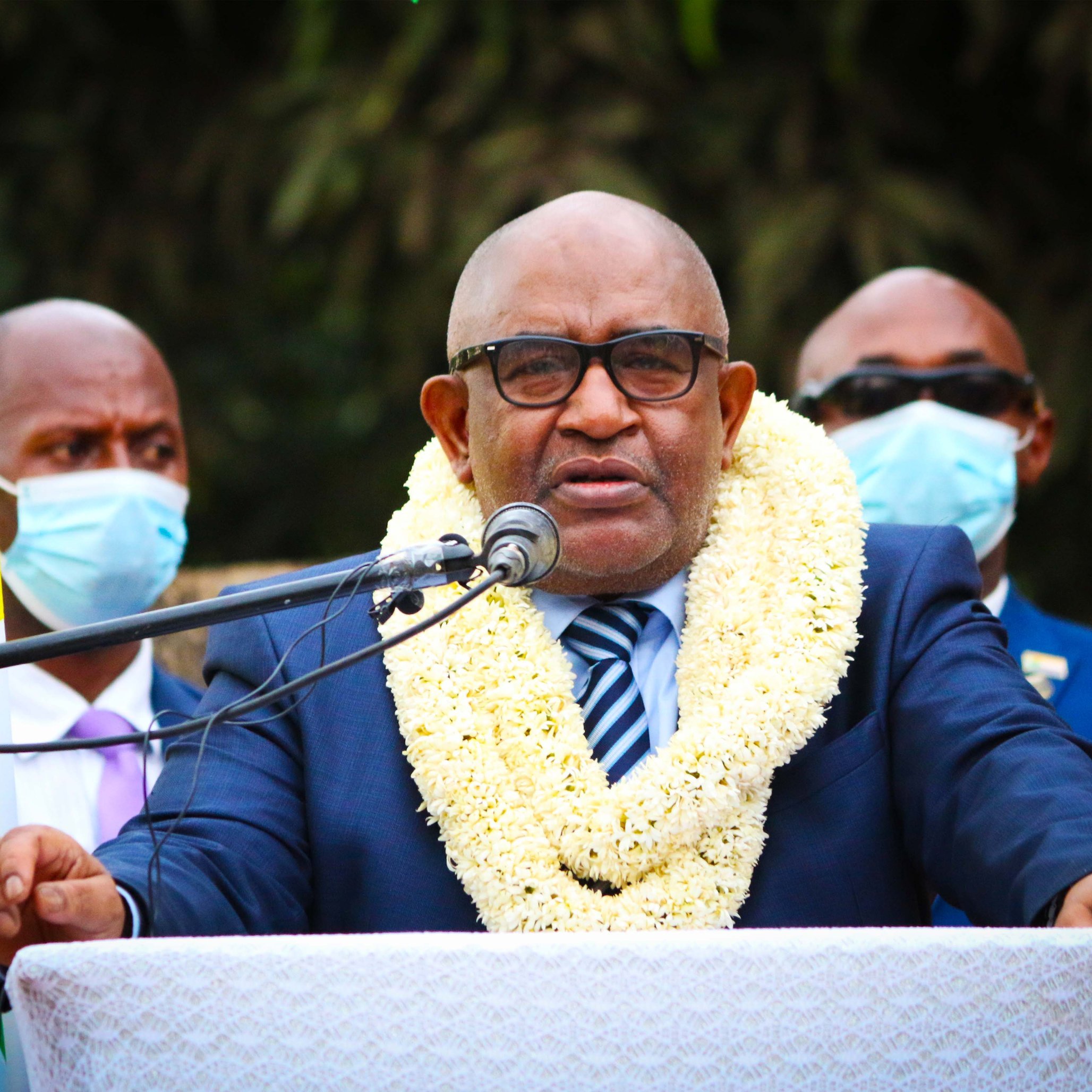 Sur cette image, un homme est debout devant un microphone, en train de s'exprimer. Il porte un costume sombre avec une chemise claire et une cravate rayée. Autour de son cou, une couronne de fleurs blanches et jaunes symbolise un accueil ou un hommage. En arrière-plan, plusieurs personnes l'écoutent attentivement, certaines portant des masques de protection. À gauche, on peut voir des drapeaux aux couleurs vives qui ajoutent une touche festive à la scène. L'ambiance semble sérieuse et solennelle.