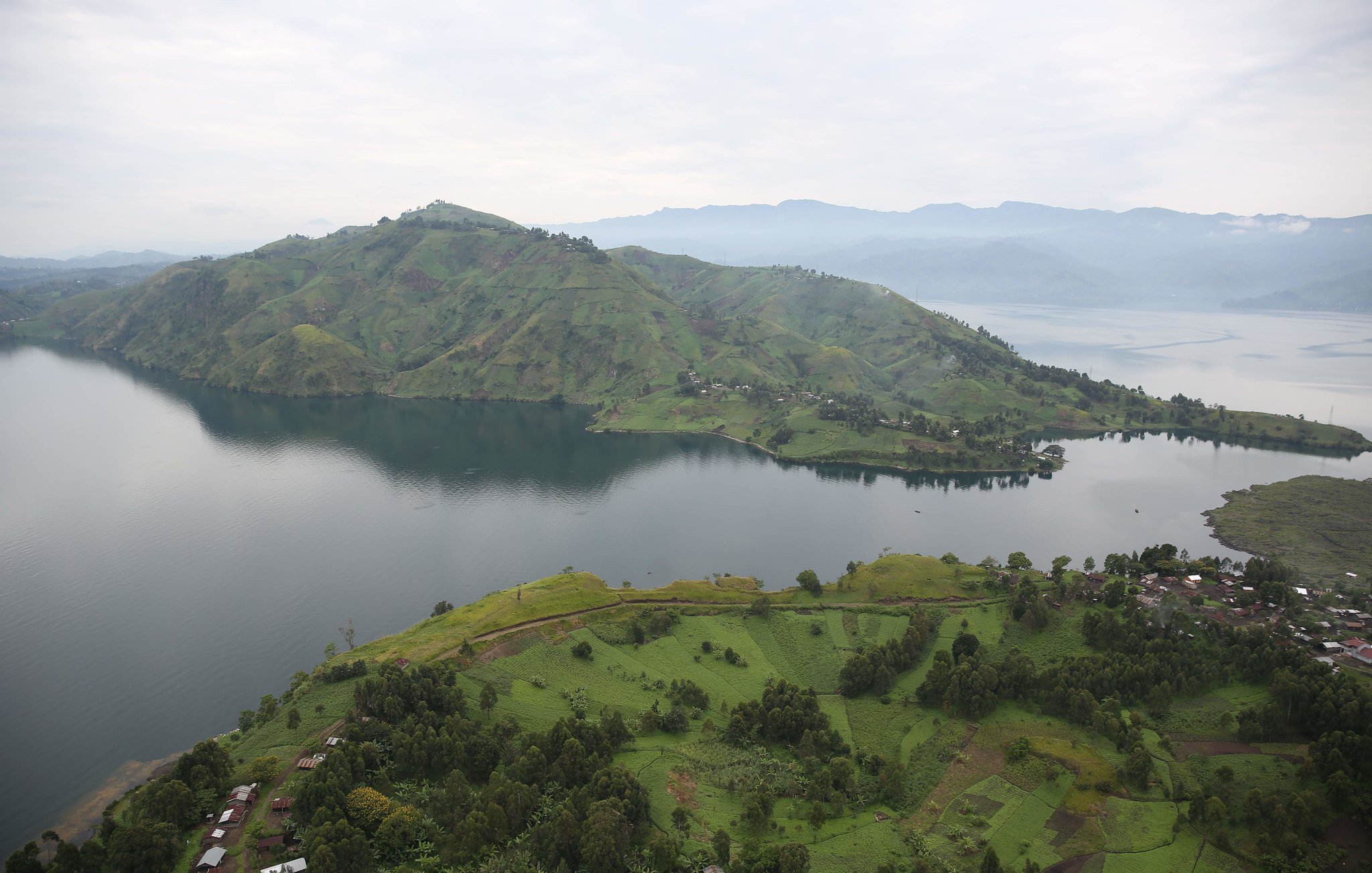 The image showcases a breathtaking landscape featuring lush green hills and a large lake. The hills are gently sloped and dotted with patches of vegetation, indicating a fertile area. In the foreground, there are scattered houses or structures, while the calm waters of the lake reflect the surrounding scenery. In the background, more mountainous terrain rises, adding depth to the view under a slightly overcast sky. The overall atmosphere is serene and picturesque, highlighting the natural beauty of the region.