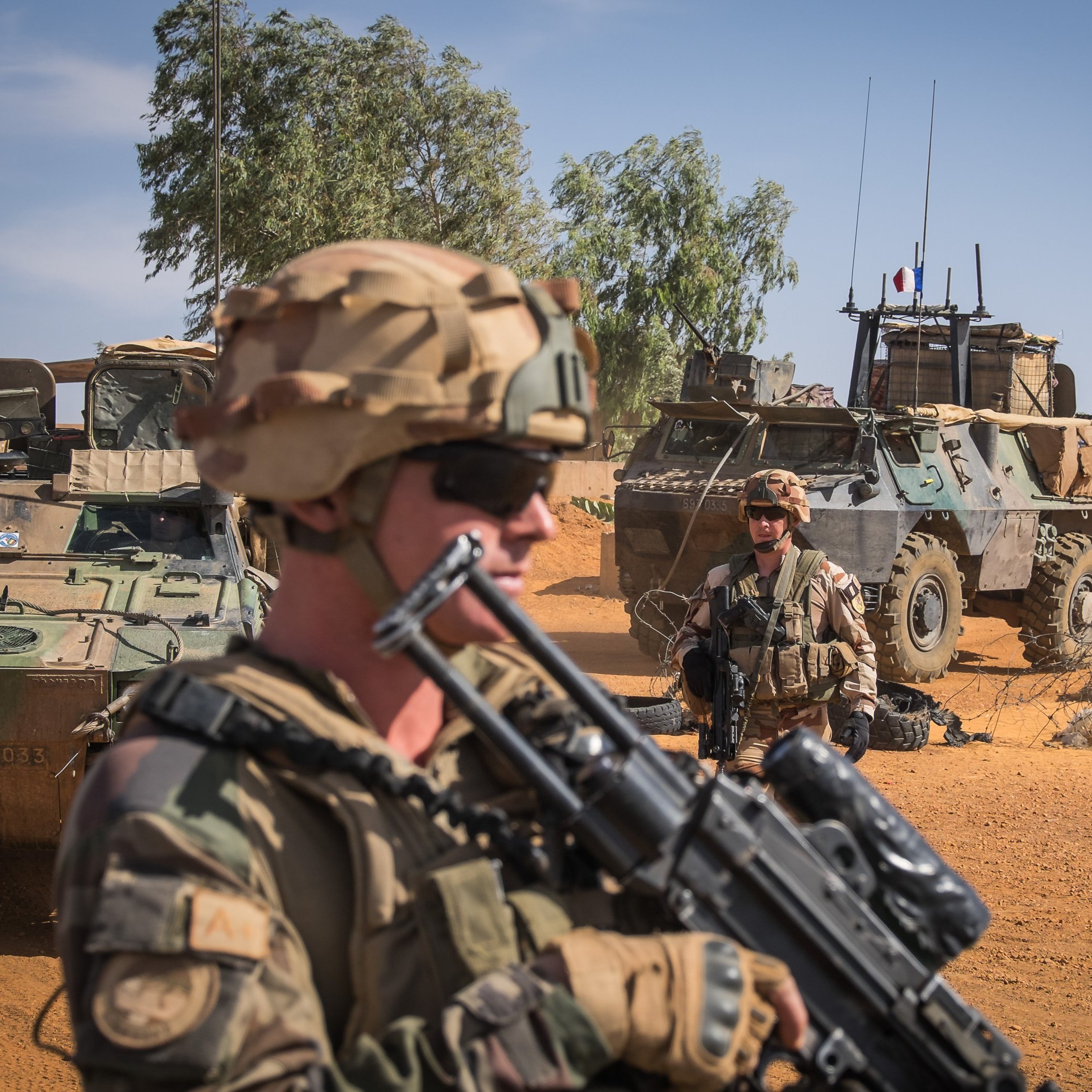 L'image montre un désert, avec un ciel bleu et quelques nuages. Au premier plan, des soldats portant des uniformes camouflés et des casques. L'un d'eux est en position de vigilance, tenant un fusil. Derrière eux, on aperçoit des véhicules militaires, dont un véhicule blindé, stationné sur un terrain sablonneux. L'environnement est aride, parsemé de quelques arbres au loin, mettant en avant une ambiance de mission militaire dans un paysage désertique.