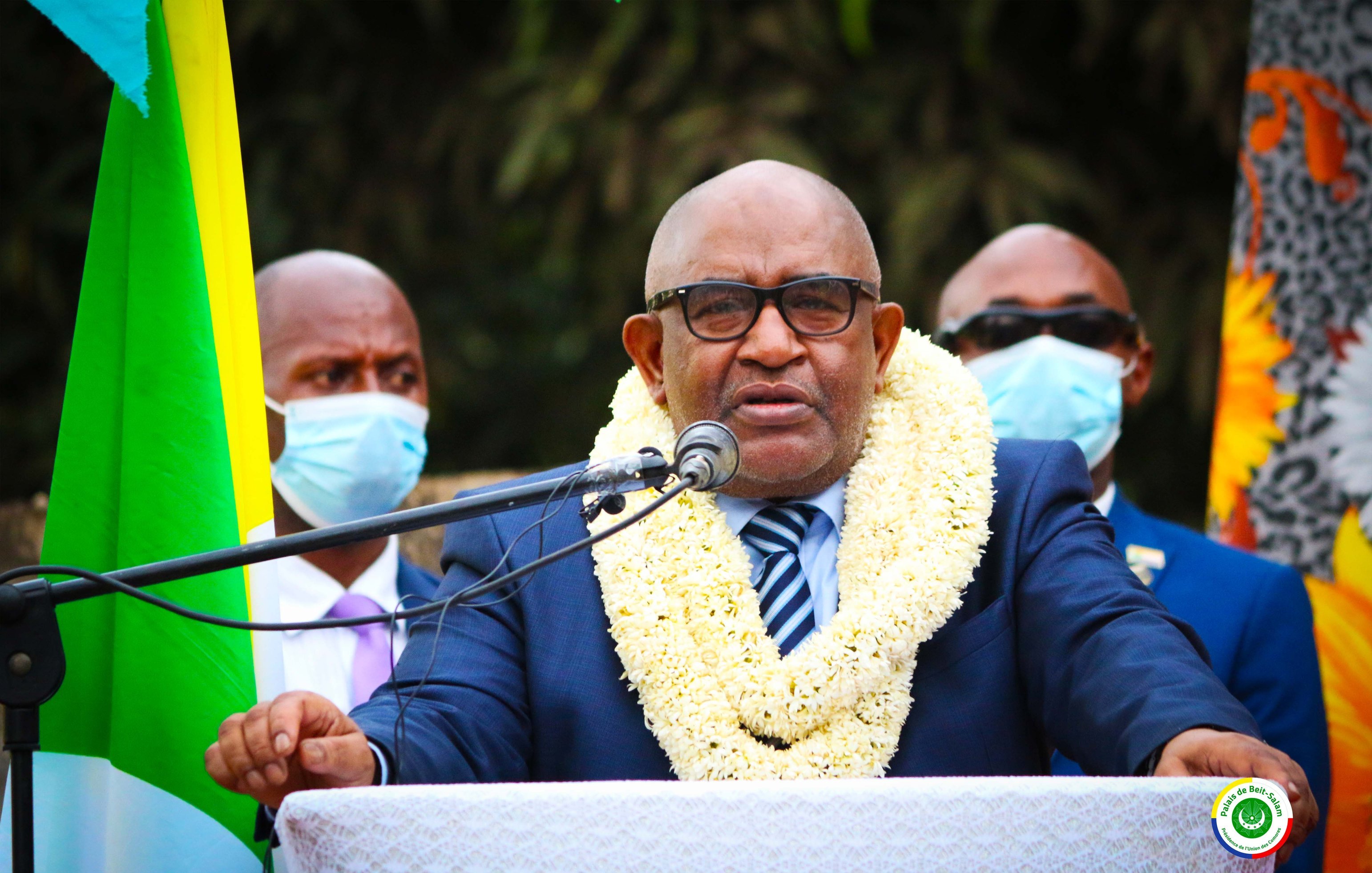 Sur cette image, un homme est debout devant un microphone, en train de s'exprimer. Il porte un costume sombre avec une chemise claire et une cravate rayée. Autour de son cou, une couronne de fleurs blanches et jaunes symbolise un accueil ou un hommage. En arrière-plan, plusieurs personnes l'écoutent attentivement, certaines portant des masques de protection. À gauche, on peut voir des drapeaux aux couleurs vives qui ajoutent une touche festive à la scène. L'ambiance semble sérieuse et solennelle.