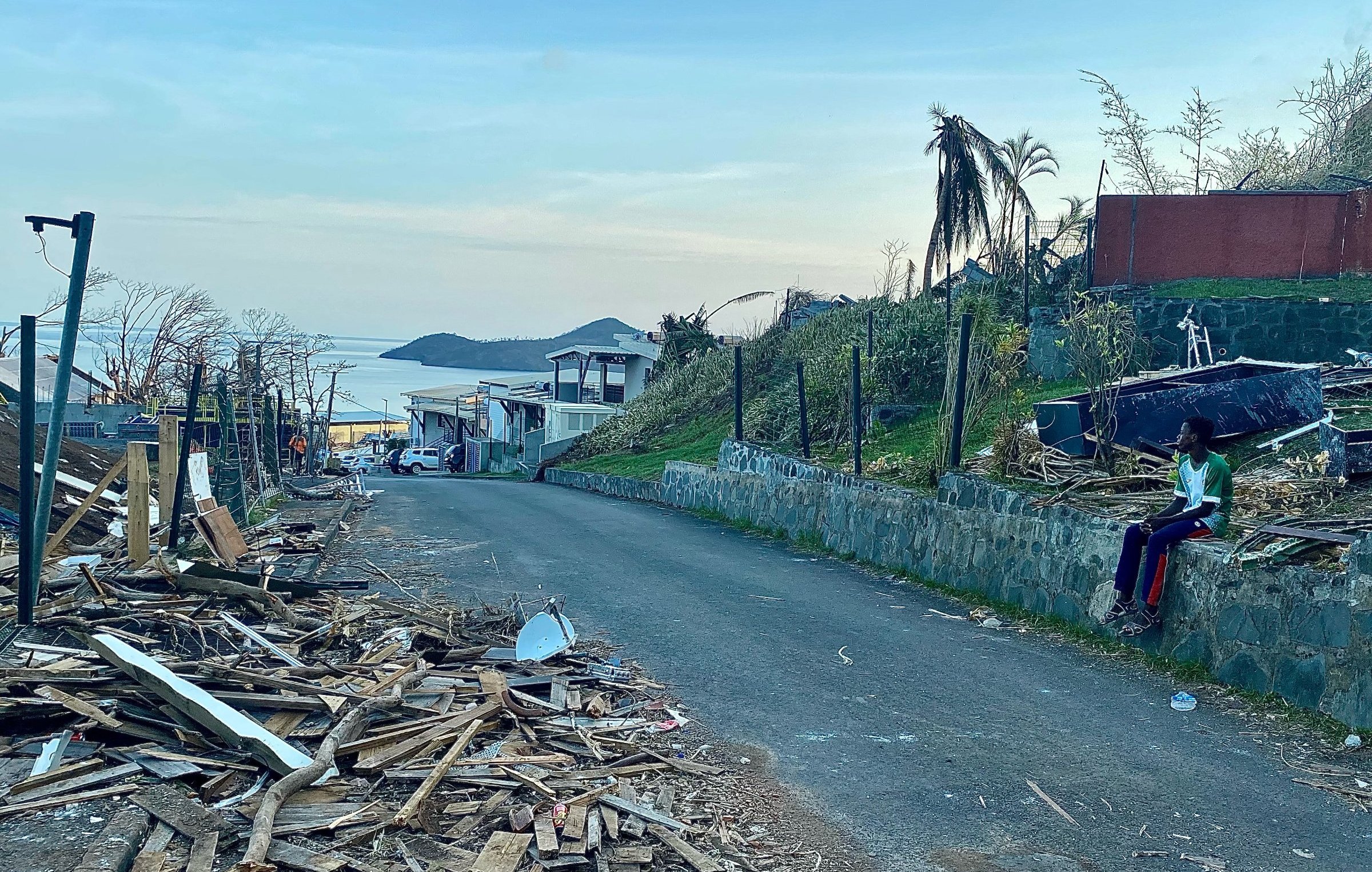 L'image présente une scène de destruction dans une rue. On voit des débris en bois éparpillés sur le sol, témoignant d'un événement dévastateur, peut-être une tempête ou un ouragan. Sur la gauche, il y a des maisons endommagées, certaines avec des toits manquants. En arrière-plan, on devine un paysage côtier avec une étendue d'eau qui reflète la lumière du ciel. À droite, une personne est assise sur un mur, habillée de vêtements colorés, semblant contempler les dégâts. La lumière du jour est douce, annonçant un crépuscule paisible, contrastant avec la morosité de la scène. Les palmiers, certains déracinés, ajoutent à l'impression de désolation.