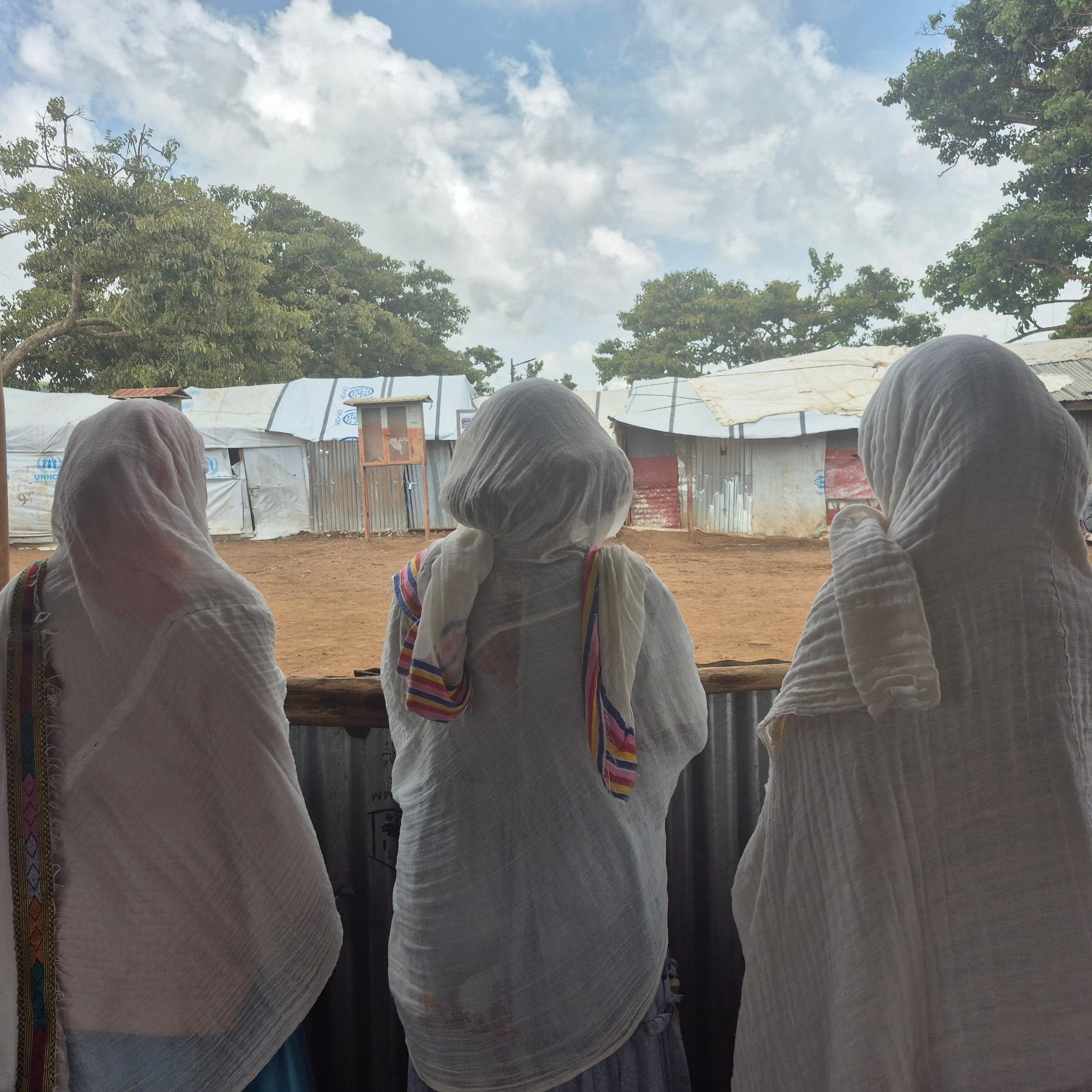 L'image montre trois femmes de dos, se tenant côte à côte, regardant à travers une structure en bois ouverte. Elles portent des vêtements longs et légers. Leurs têtes sont couvertes de foulards, ce qui suggère un contexte culturel ou religieux. Au loin, on aperçoit des abris temporaires, probablement des tentes ou des structures en tôles. Le ciel est partiellement nuageux, créant une atmosphère paisible. L'environnement évoque un lieu de rassemblement ou d'attente, avec un sol en terre battue. La scène semble calme, laissant entrevoir un moment de contemplation ou de partage entre ces femmes.