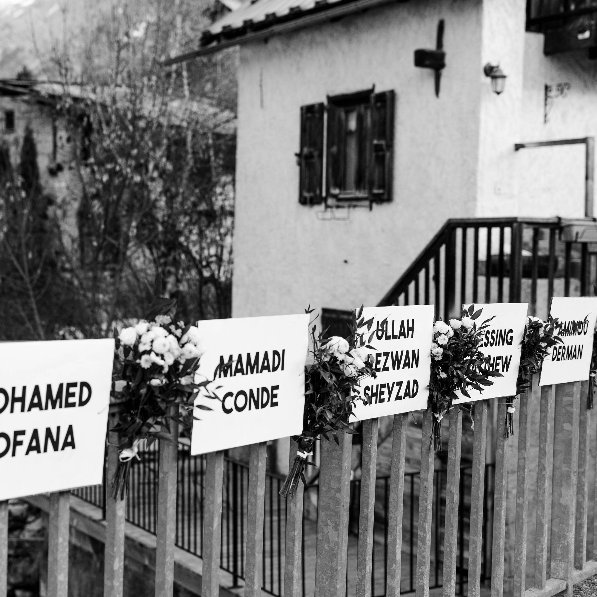 L'image montre une scène en noir et blanc, où l'on aperçoit une maison avec un balcon en bois. Devant la maison, il y a une clôture sur laquelle sont fixés plusieurs panneaux blancs. Chaque panneau présente un prénom et un nom, probablement en hommage à des personnes. Des fleurs blanches sont disposées autour de chaque panneau, ajoutant une touche de douceur. En arrière-plan, on peut deviner des arbres et la nature environnante, suggérant un cadre paisible et naturel.