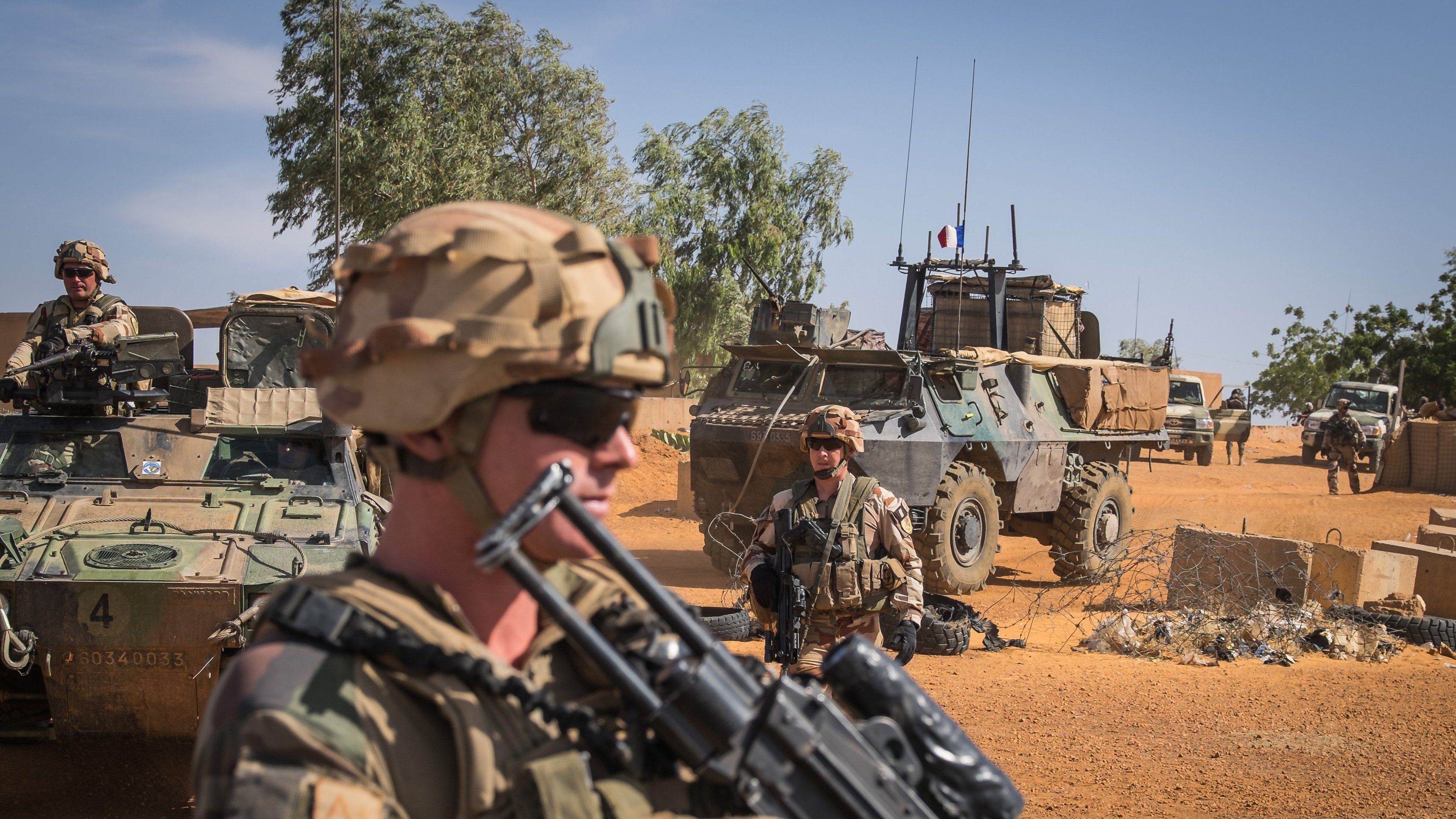 L'image montre un paysage désertique, typique d'une zone militaire. Au premier plan, un soldat est en position, tenant un fusil. Il porte un casque et des lunettes de soleil, ce qui lui confère une allure déterminée. En arrière-plan, on aperçoit d'autres soldats et un véhicule blindé, avec des éléments de terrain militaire, comme des barbelés et des équipements. Le ciel est dégagé, et la lumière du soleil éclaire la scène, soulignant l'environnement aride et la présence militaire. L'atmosphère respire la vigilance et la préparation.