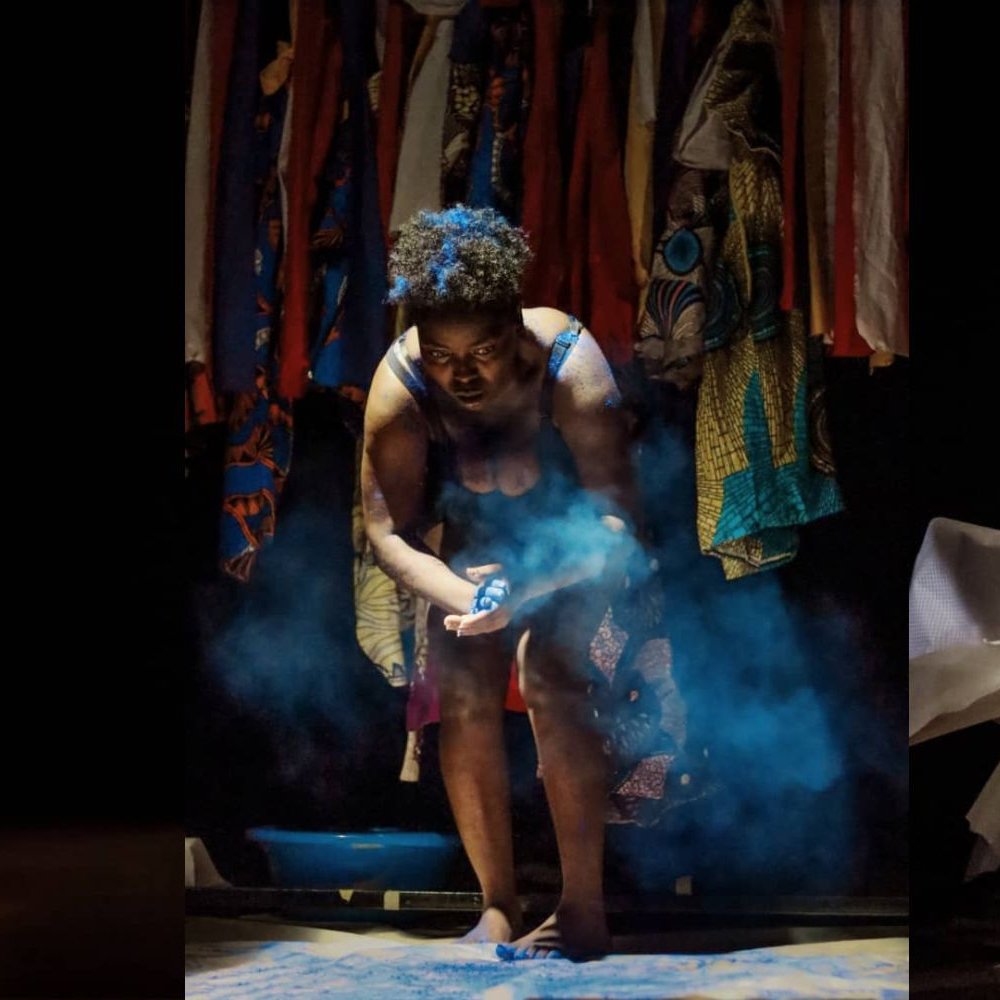 L'image présente une scène de performance artistique en trois parties. À gauche, on voit une silhouette sombre d'une danseuse, légèrement éclairée, qui se tient debout, les jambes nues, suggérant une posture pensive ou introspective. Au centre, une autre danseuse est en action, entourée de fumée, en train de manipuler des objets, évoquant une intensité émotionnelle et une interaction avec l'espace. Enfin, à droite, la danseuse semble déchirer des morceaux de tissu, exprimant un mouvement dynamique et presque chaotique. En arrière-plan, une tapisserie colorée de tissus suspendus ajoute une texture visuelle riche et un contraste vibrant à la scène sombre. L'ensemble crée une atmosphère à la fois intime et expressive, transmettant des émotions variées à travers le mouvement et la présence corporelle.