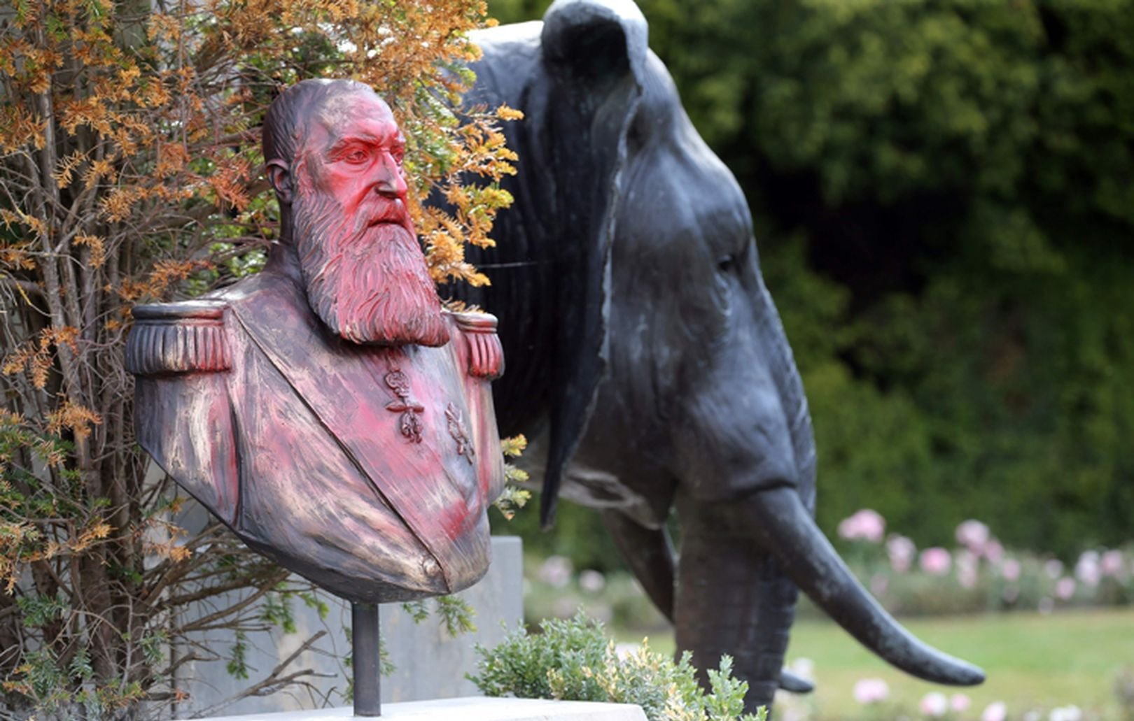 L'image présente un buste en bronze d'un homme barbu, aux cheveux longs et portant des uniformes militaires, posé sur un socle. Ce buste est entouré de végétation, ce qui lui donne un aspect naturel. En arrière-plan, on aperçoit la silhouette d'un éléphant en métal, affichant des détails réalistes comme ses grandes oreilles et sa trompe. Le contraste entre le buste humain et l'éléphant, ainsi que la verdure environnante, crée une ambiance paisible et mémorable dans un jardin.