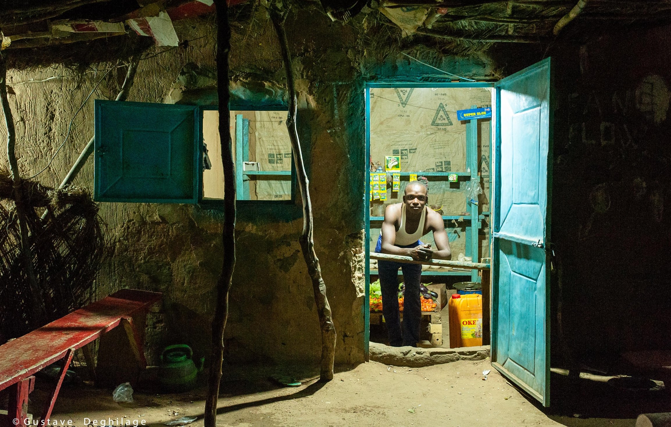 L'image montre l'entrée d'une petite boutique, éclairée dans l'obscurité. La porte est ouverte, révélant un intérieur modeste. On aperçoit un homme qui se tient derrière un comptoir, son regard tourné vers l'extérieur. Il porte un débardeur et semble attentif, probablement en attente de clients. Les murs sont en terre, et des papiers et affiches sont accrochés, ajoutant une touche de couleur à l'environnement. À l'avant, un banc en bois rouge est installé, et quelques bouteilles et contenants sont visibles à l'intérieur de la boutique. L'ambiance est chaleureuse malgré le contexte rural et simple.