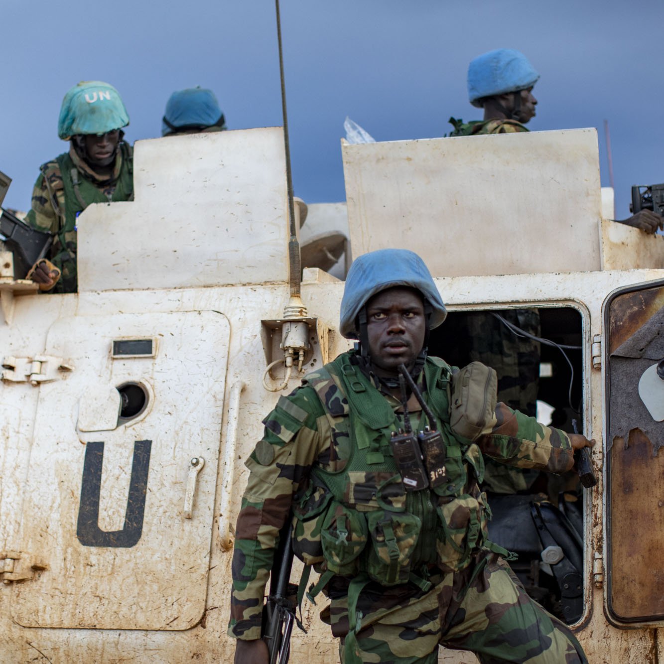 L'image montre un groupe de soldats en uniforme, portant des casques bleus emblématiques des Casques bleus de l'ONU. Ils sont près d'un véhicule militaire, sale et marqué par la boue, ce qui indique des conditions difficiles. Le soldat en premier plan, avec un regard déterminé, semble descendre du véhicule. À l'arrière, d'autres soldats se tiennent, prêts à intervenir. Le ciel est nuageux, ajoutant une ambiance sombre et sérieuse à la scène, qui évoque une mission de maintien de la paix dans un environnement hostile.