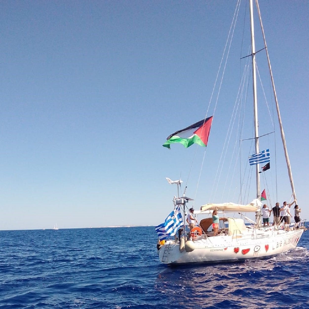 The image depicts a sailboat on calm blue waters. The boat features several flags flying from its mast: notably, a Palestinian flag, a Greek flag, and a blue-and-white striped flag typical of Greece. The sky is clear and bright, suggesting a sunny day at sea. The surrounding water appears serene, reflecting the sunlight.