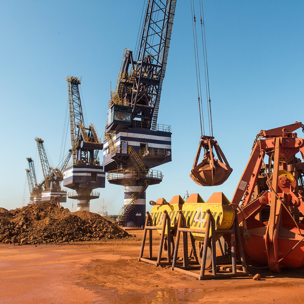 Dans cette image, nous sommes dans un port industriel où plusieurs grues sont visibles. Ces grandes machines sont utilisées pour charger et décharger des matériaux. À l'avant, il y a des godets et des bennes orange, qui sont des équipements de levage, reposant sur le sol. En arrière-plan, plusieurs grues se dressent contre un ciel bleu clair, leurs bras et leurs crochets descendus vers les tas de terre brune. L'atmosphère est typiquement industrielle, avec un sol en terre battue et une ambiance de travail intense. Des sons de machines en fonctionnement et le bruit des cargaisons en mouvement pourraient être présents dans cet environnement.