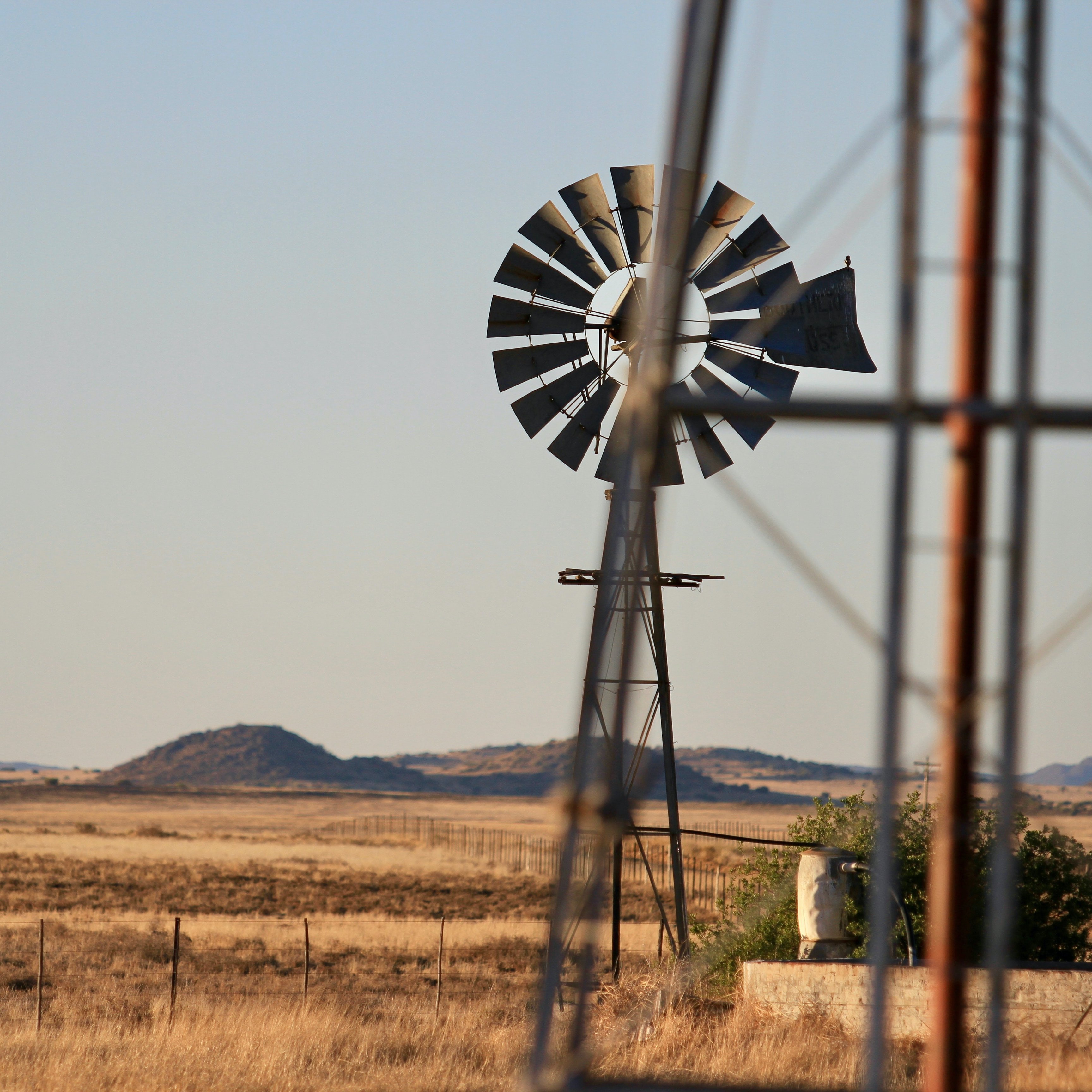 L'image montre un paysage rural étendu sous un ciel dégagé. À l'arrière-plan, des collines douces se dessinent sur l'horizon, tandis que le sol est recouvert d'une herbe sèche, témoignant d'un environnement aride. Au premier plan, une éolienne en métal tourne doucement, projetant son ombre sur le sol. Les pales de l'éolienne sont bien visibles, capturant quelques rayons de soleil. À proximité, un pilier métallique se dresse légèrement flou, ajoutant une touche de profondeur à la scène. L'ensemble évoque une atmosphère calme et tranquille, comme un moment suspendu dans la nature.