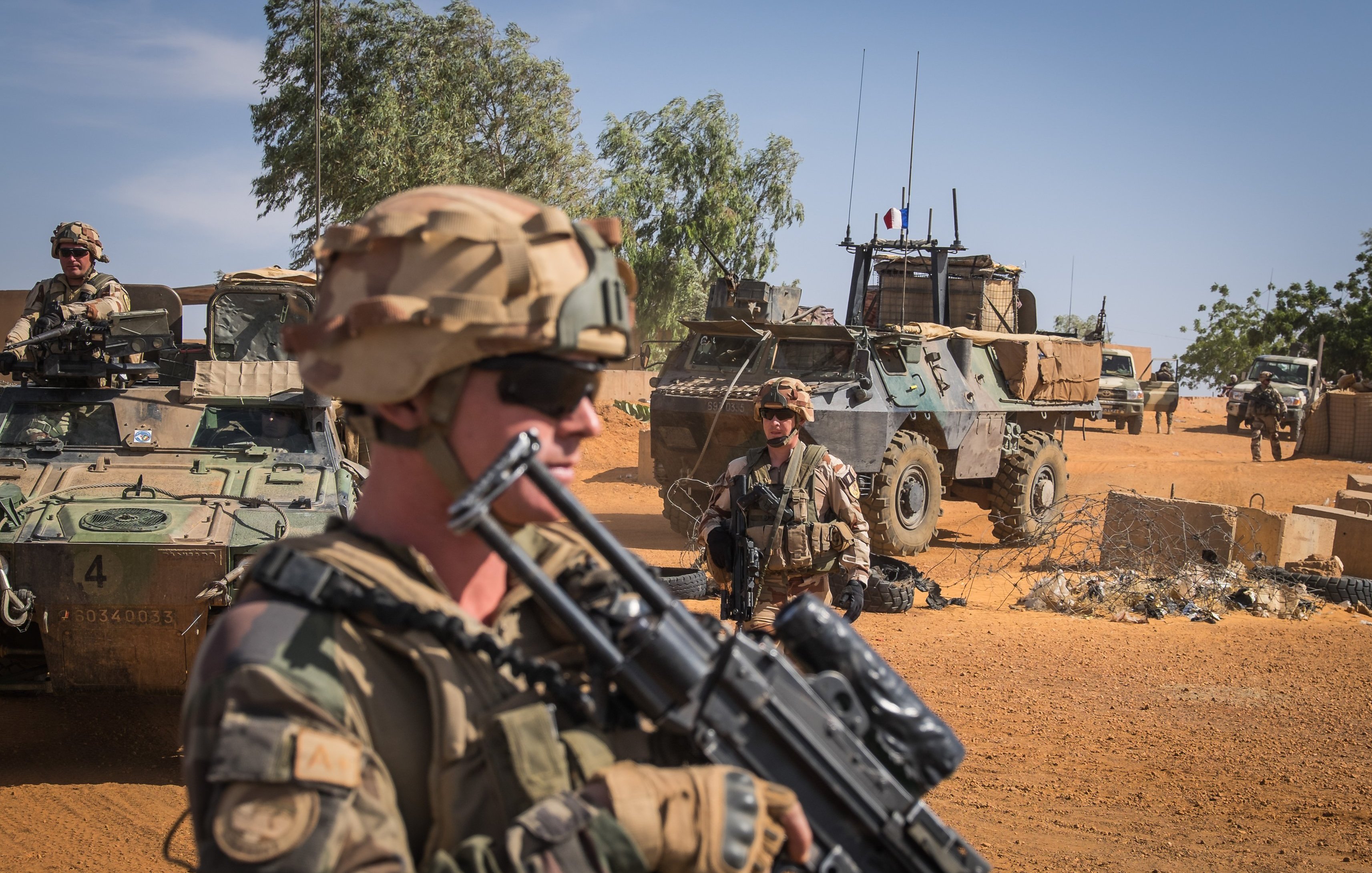 L'image montre un désert, avec un ciel bleu et quelques nuages. Au premier plan, des soldats portant des uniformes camouflés et des casques. L'un d'eux est en position de vigilance, tenant un fusil. Derrière eux, on aperçoit des véhicules militaires, dont un véhicule blindé, stationné sur un terrain sablonneux. L'environnement est aride, parsemé de quelques arbres au loin, mettant en avant une ambiance de mission militaire dans un paysage désertique.