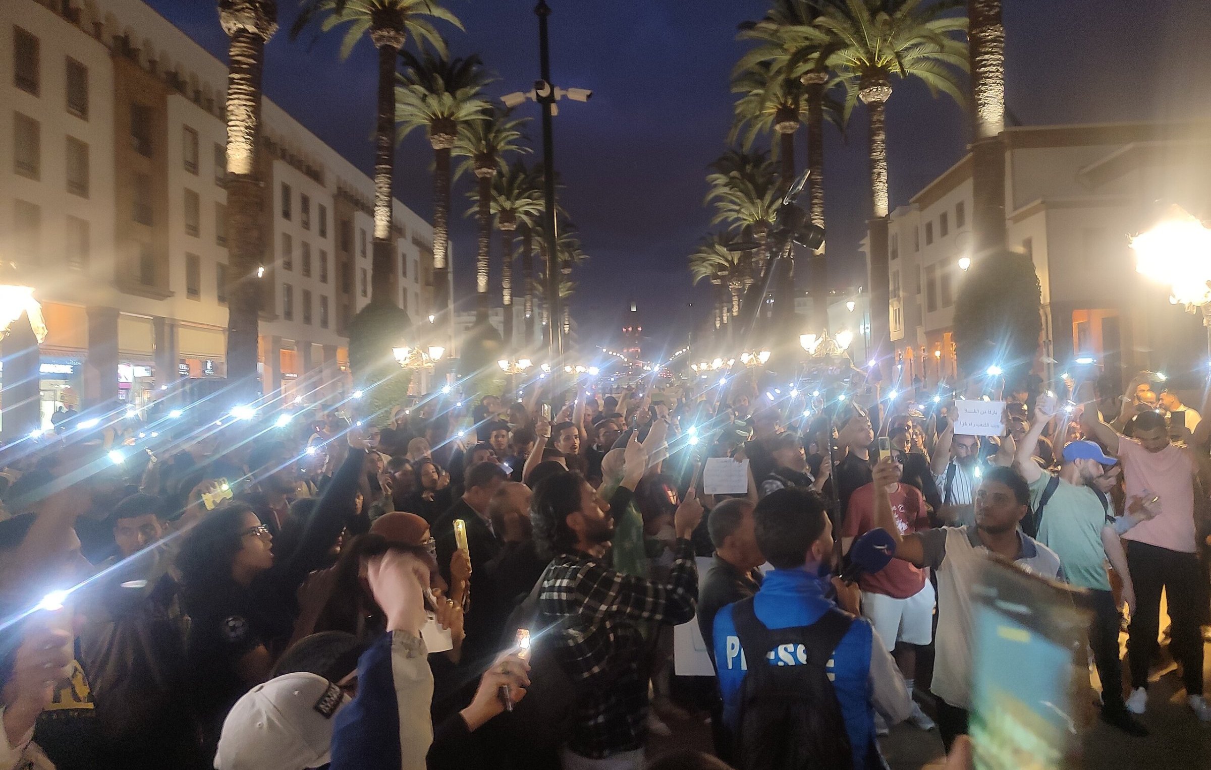 L'image montre une grande foule rassemblée dans une rue animée, éclairée par des lampadaires et entourée de palmiers. La scène se déroule en soirée, avec un ciel crépusculaire. Beaucoup de personnes tiennent des téléphones ou des lumières, créant un effet lumineux impressionnant. Les gens semblent mobilisés, certains tenant des pancartes et s'exprimant avec passion. L'atmosphère est énergique et engagée, reflétant une ambiance de manifestation ou d'événement public où les participants cherchent à faire entendre leur voix.