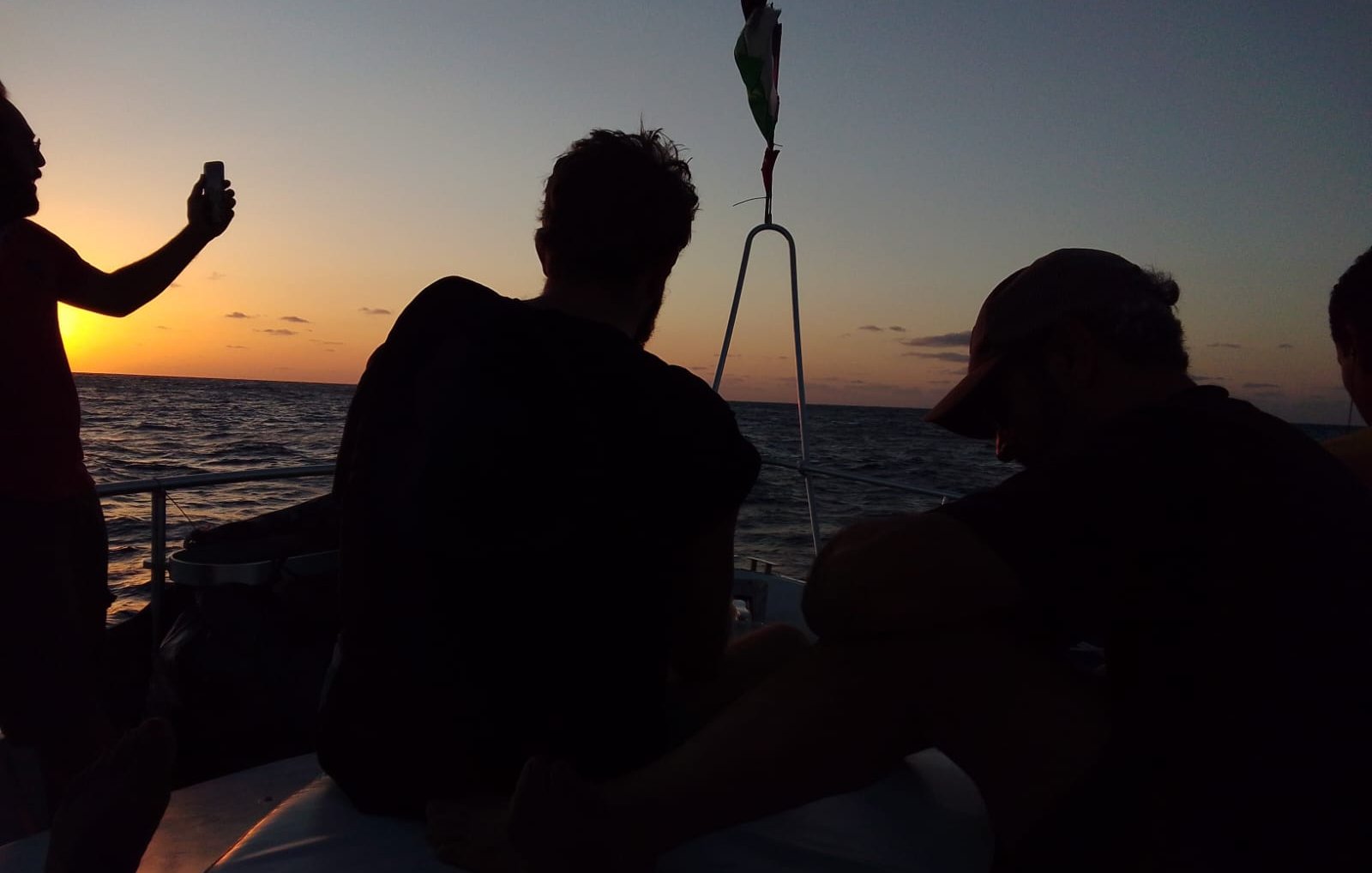 The image shows a group of people on a boat at sunset. The sky is painted in warm hues of orange and yellow as the sun sets on the horizon. Two people are visible, sitting with their backs to the camera, seemingly enjoying the view. One person is standing and holding a phone, possibly taking a photo. The water appears calm, reflecting the colors of the sky. The overall atmosphere is serene and relaxed.