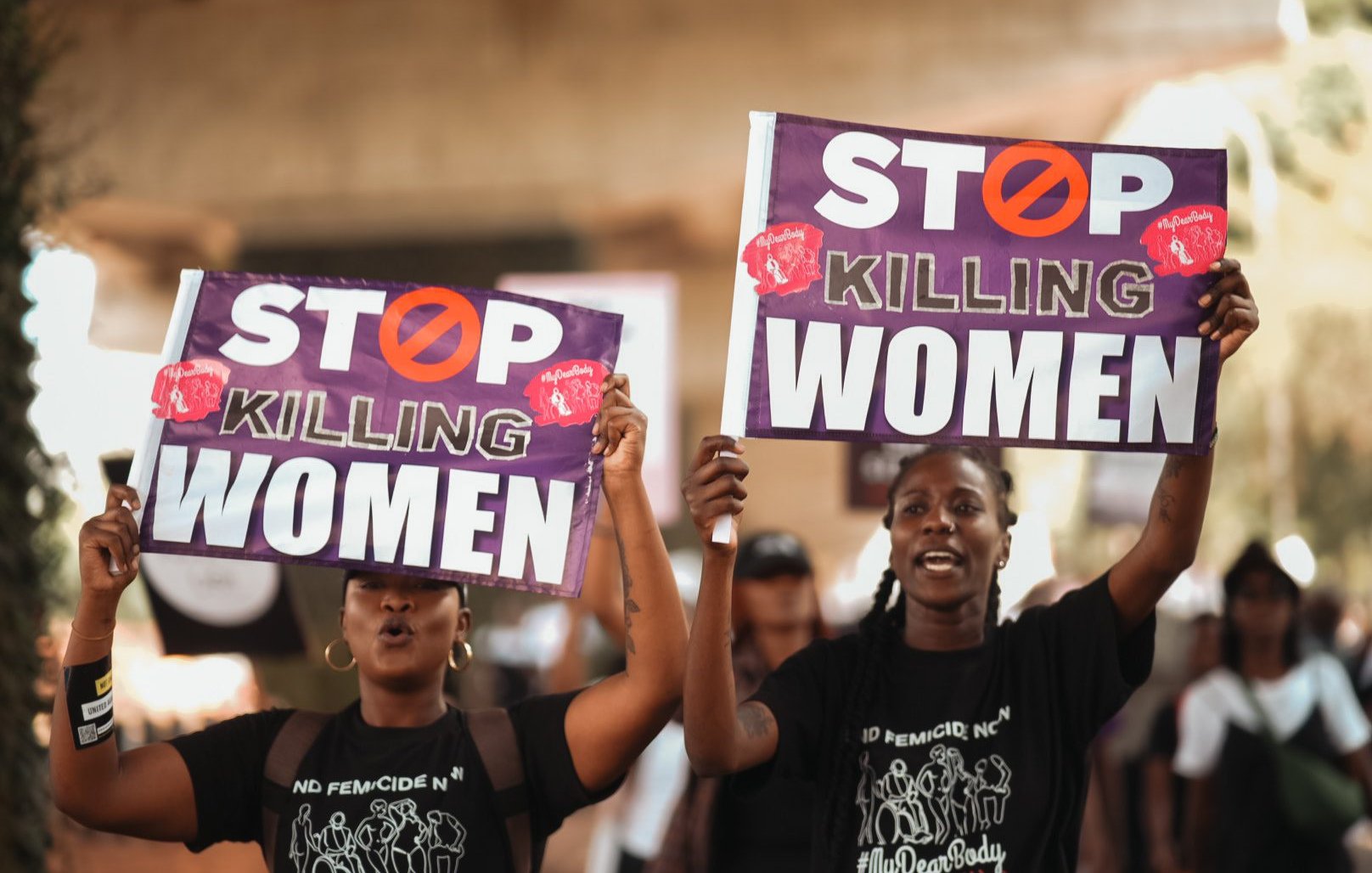 L'image montre deux femmes participante à une manifestation. Elles tiennent chacune une grande pancarte. Les pancartes portent le message "STOP KILLING WOMEN" en lettres capitales de couleur blanche, avec un symbole barré en rouge. Les femmes ont des expressions déterminées et sont vêtues de t-shirts noirs. L'arrière-plan suggère une ambiance de mobilisation avec d'autres personnes qui marchent et participent à l'événement. L'atmosphère est chargée d'émotion et de solidarité.