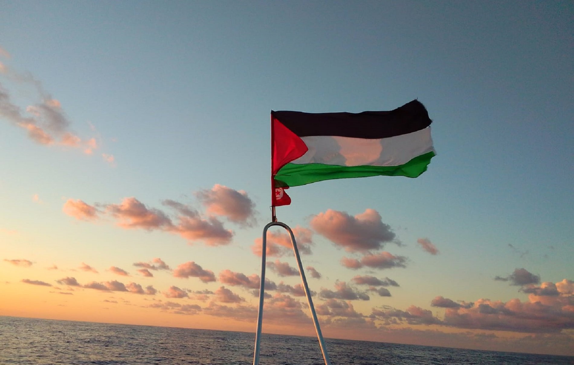 L'image montre un drapeau palestinien flottant sur un mât, avec un vaste horizon marin au fond. Le ciel est teinté de couleurs chaudes, probablement à l'aube ou au crépuscule, avec des nuages épars. Les couleurs du drapeau sont distinctes : le noir, le blanc, le vert et le rouge. La mer est calme et reflète doucement les teintes du ciel, créant une ambiance paisible et solennelle.