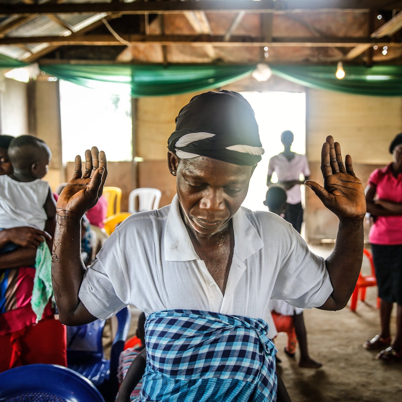 L'image montre un intérieur de salle, où des chaises colorées sont disposées de manière informelle. Au premier plan, une femme se tient debout, les bras levés, dans une posture de prière ou de méditation. Elle porte un foulard sur la tête et une blouse blanche, tandis qu'une jupe à motifs colorés entoure sa taille. En arrière-plan, on aperçoit d'autres personnes, certaines sont engagées dans une conversation tandis que d'autres semblent également en prière. L'ambiance est empreinte de spiritualité et d'un sentiment de communauté. La lumière naturelle entre par des fenêtres, créant un espace à la fois chaleureux et intime.