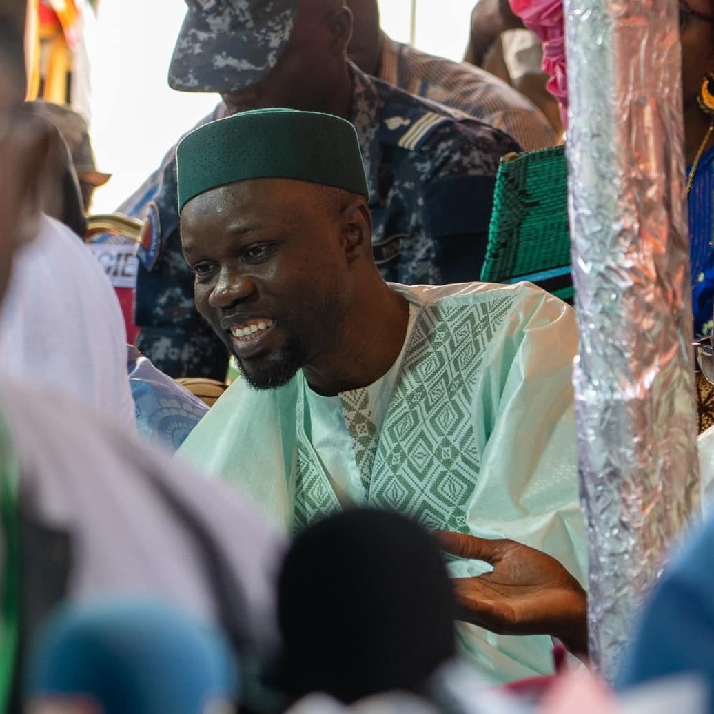 L'image montre un groupe de personnes rassemblées dans un environnement animé, probablement lors d'un événement public. Au premier plan, un homme souriant est vêtu d'un habit traditionnel, avec un turban ou une coiffe verte. Il semble engagé dans une discussion, ses mains gesticulant pour accompagner ses paroles. En arrière-plan, d'autres personnes sont présentes, certaines en uniforme, suggérant un contexte formel ou officiel. La scène est remplie de couleurs vives, et l'atmosphère semble dynamique et interactive.