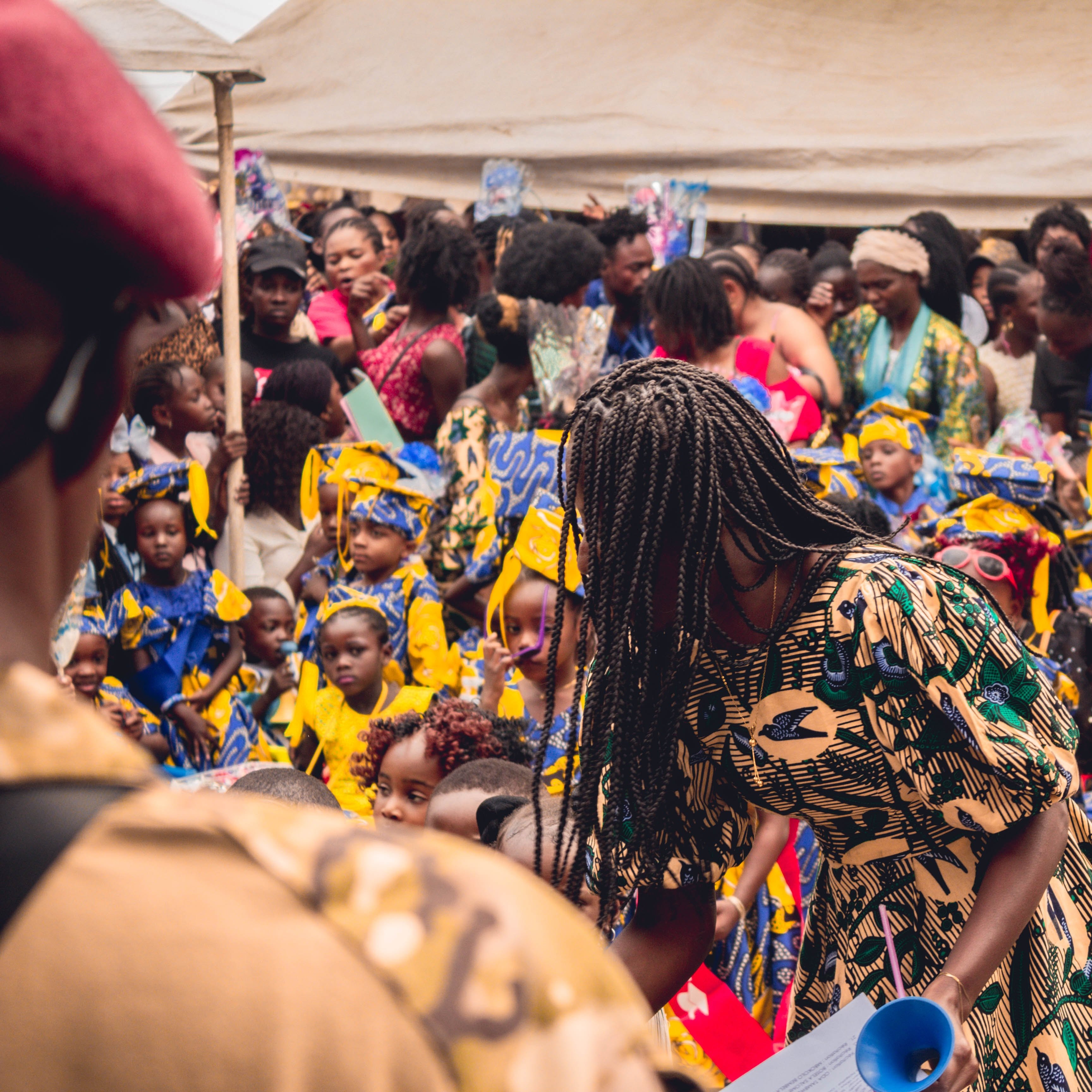L'image montre une scène vibrante et animée d'un événement en plein air, probablement une célébration ou un festival. Au premier plan, on voit une femme portant une robe aux motifs colorés qui se penche, en train de distribuer quelque chose. Derrière elle, un grand groupe de filles, habillées de robes jaunes ornées de motifs bleus, se tient avec des sourires sur le visage. Leur ambiance semble joyeuse et festive. La foule est dense, avec de nombreuses personnes, y compris des femmes et des enfants, qui interagissent et profitent de l'événement. Des éléments décoratifs, comme des ballons ou des banderoles, pourraient ajouter une touche festive à l'environnement. L'atmosphère globale dégage une sensation de communauté et de célébration.
