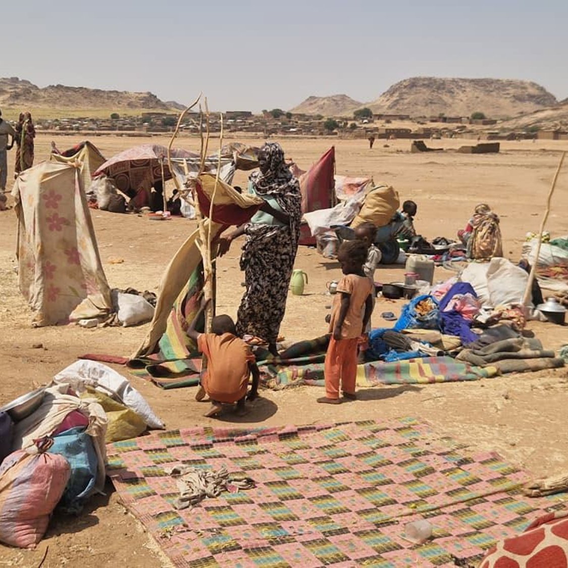 L'image montre un campement dans une région aride et désertique. On peut imaginer une vaste étendue de terrain sec, avec quelques collines en arrière-plan. Des tentes rudimentaires, faites de toile ou de vêtements usagés, sont disposées de manière désordonnée, offrant un semblant de refuge aux personnes qui y vivent. Au sol, des tapis ou des morceaux de tissu sont étalés, tandis que des objets personnels, comme des sacs et des casseroles, sont éparpillés autour. Au centre de la scène, un groupe de personnes est en train de s'activer : une femme semble organiser quelque chose, tandis qu'un enfant debout à côté d'elle observe. L'atmosphère transpire la précarité mais aussi une certaine résilience face aux conditions difficiles. Les couleurs des tissus sont variées, mais plutôt ternes et fanées par le temps. L'ensemble dégage une impression de lutte pour la survie dans un environnement hostile.