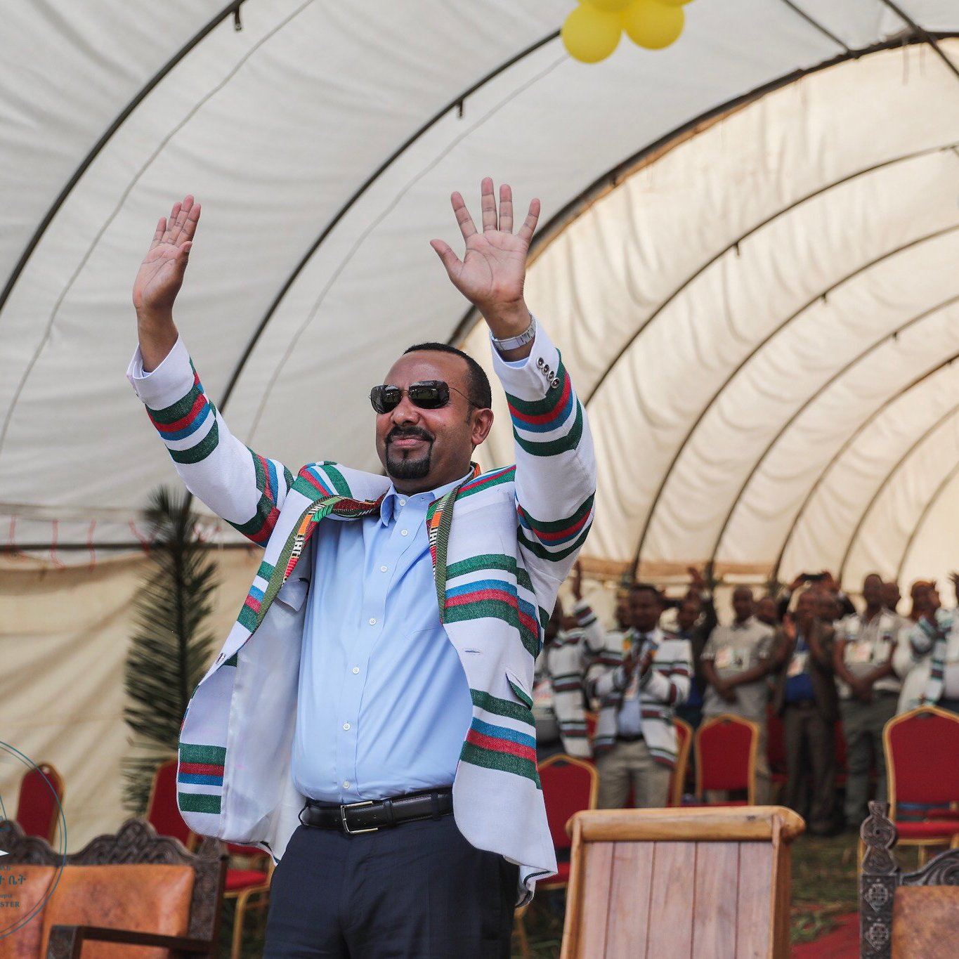 L'image montre un homme, probablement un leader, se tenant debout sous un grand chapiteau. Il porte une tenue colorée avec des rayures multicolores et des lunettes de soleil, et lève les deux mains dans un geste de salutation ou d'encouragement. En arrière-plan, on aperçoit un groupe de personnes qui semblent l'écouter attentivement, tous habillés de manière décontractée. Le chapiteau est décoré avec des ballons, ajoutant une ambiance festive à l'événement. L'atmosphère générale est celle d'un rassemblement joyeux et engageant.