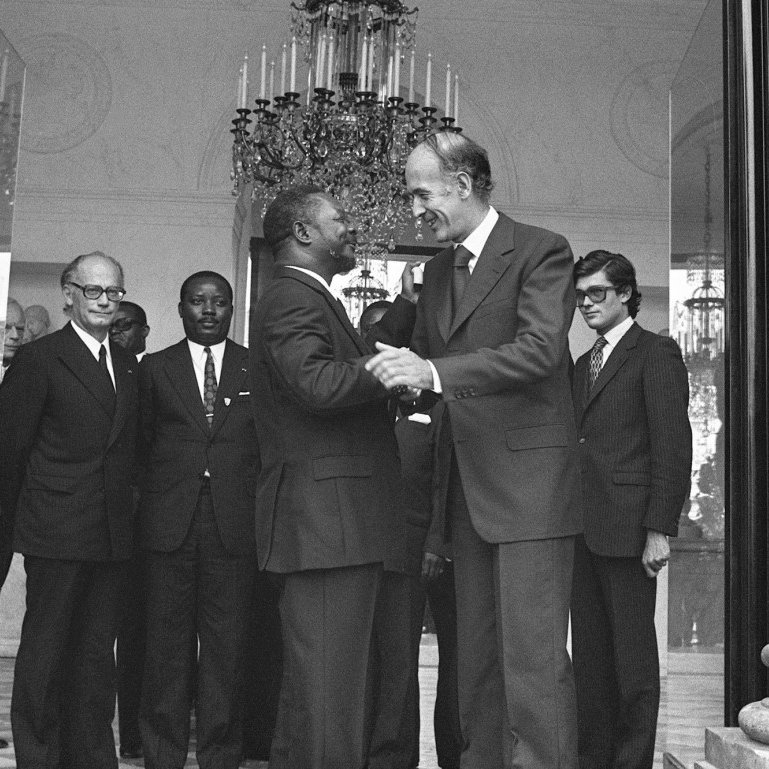 L'image montre un groupe d'hommes en costume, réunis dans un grand hall bien éclairé. Au centre, deux hommes se serrent la main, affichant des expressions de cordialité. L'un est légèrement plus âgé, avec des cheveux grisonnants, tandis que l'autre est plus jeune. En arrière-plan, plusieurs autres hommes les observent, tous vêtus de costumes sombres, ce qui accentue l'atmosphère formelle de la rencontre. Le décor est élégant, avec un grand lustre suspendu au plafond et des colonnes en pierre qui ajoutent une touche de sophistication.