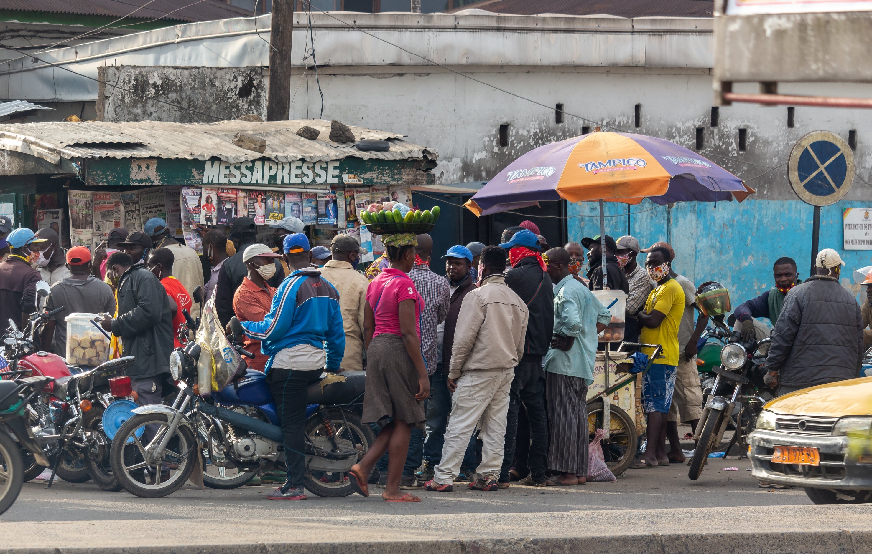 L'image montre une scène animée dans une rue d'une ville. On peut voir un groupe de personnes rassemblées autour d'un kiosque à journaux, qui semble être un lieu de rencontre. Certaines personnes portent des masques, ce qui indique une attention à la santé publique. À l'avant, une femme porte un grand panier de bananes sur sa tête, ce qui met en valeur une pratique courante dans de nombreuses cultures. Des motos et une voiture jaune sont garées à proximité, ajoutant à l'ambiance urbaine. En arrière-plan, on peut distinguer des bâtiments simples, avec des couleurs plutôt ternes, ce qui donne un aspect de vie quotidienne dans ce quartier.