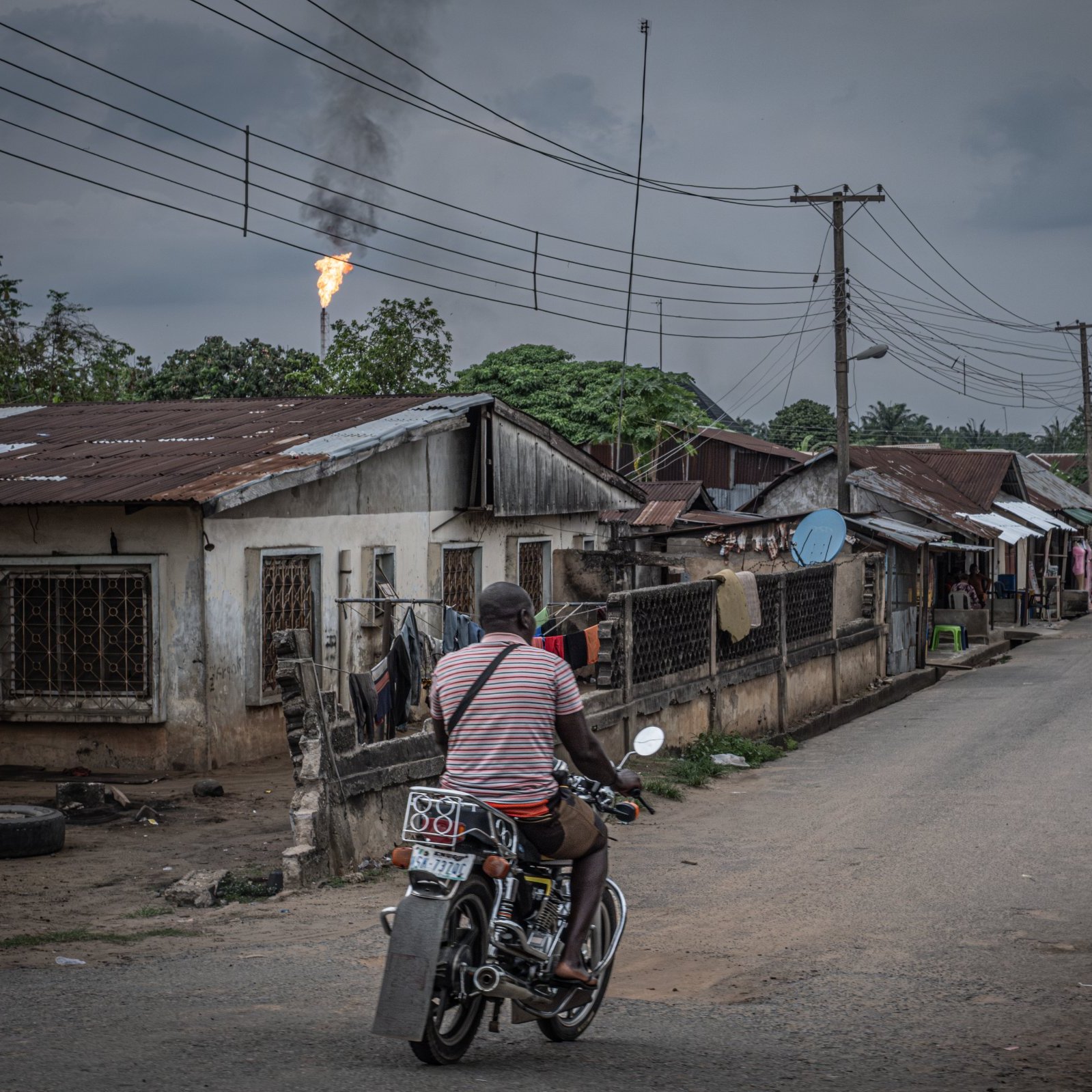 L'image présente une scène urbaine dans un quartier qui semble rural. Une route centrale est bordée de maisons modestes, souvent en tôle, avec des fenêtres grillagées. À gauche, on apperçoit un homme sur une moto, portant un t-shirt rayé, qui se dirige vers l'avant, ajoutant une touche de mouvement à la scène. En arrière-plan, des poteaux électriques sont visibles, ainsi qu'un tuyau de gaz qui rejette une flamme, indiquant peut-être une installation industrielle à proximité. Le ciel est couvert, avec des nuages sombres, créant une atmosphère légèrement mélancolique. À droite, des plantations de bananiers ajoutent une touche de verdure. L'ensemble dépeint une réalité de vie quotidienne dans un environnement à la fois simple et intéressant.
