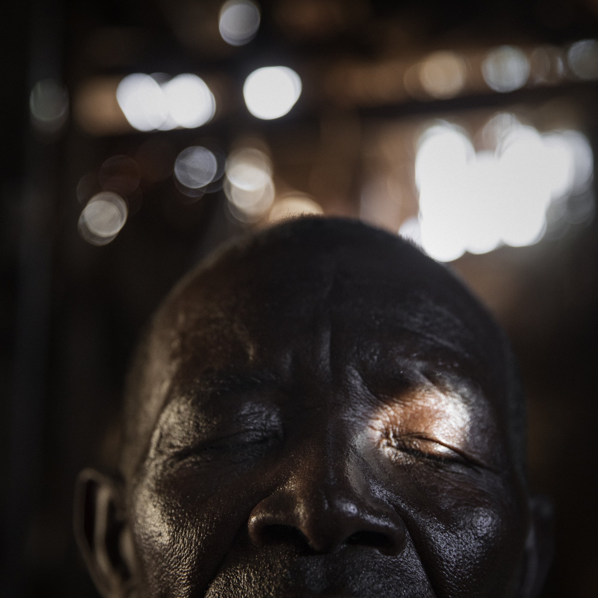 L'image montre un homme avec les yeux fermés, laissant transparaître une profonde sérénité. Son visage, marqué par le temps, présente des rides et une peau sombre. Une lumière douce filtre à travers un espace, touchant délicatement son visage et créant des reflets subtilement brillants en arrière-plan. L'ambiance de l'image est calme, évoquant un moment de méditation ou de paix intérieure. Les contours flous de l'environnement suggèrent un espace simple, sans détails distracteurs.