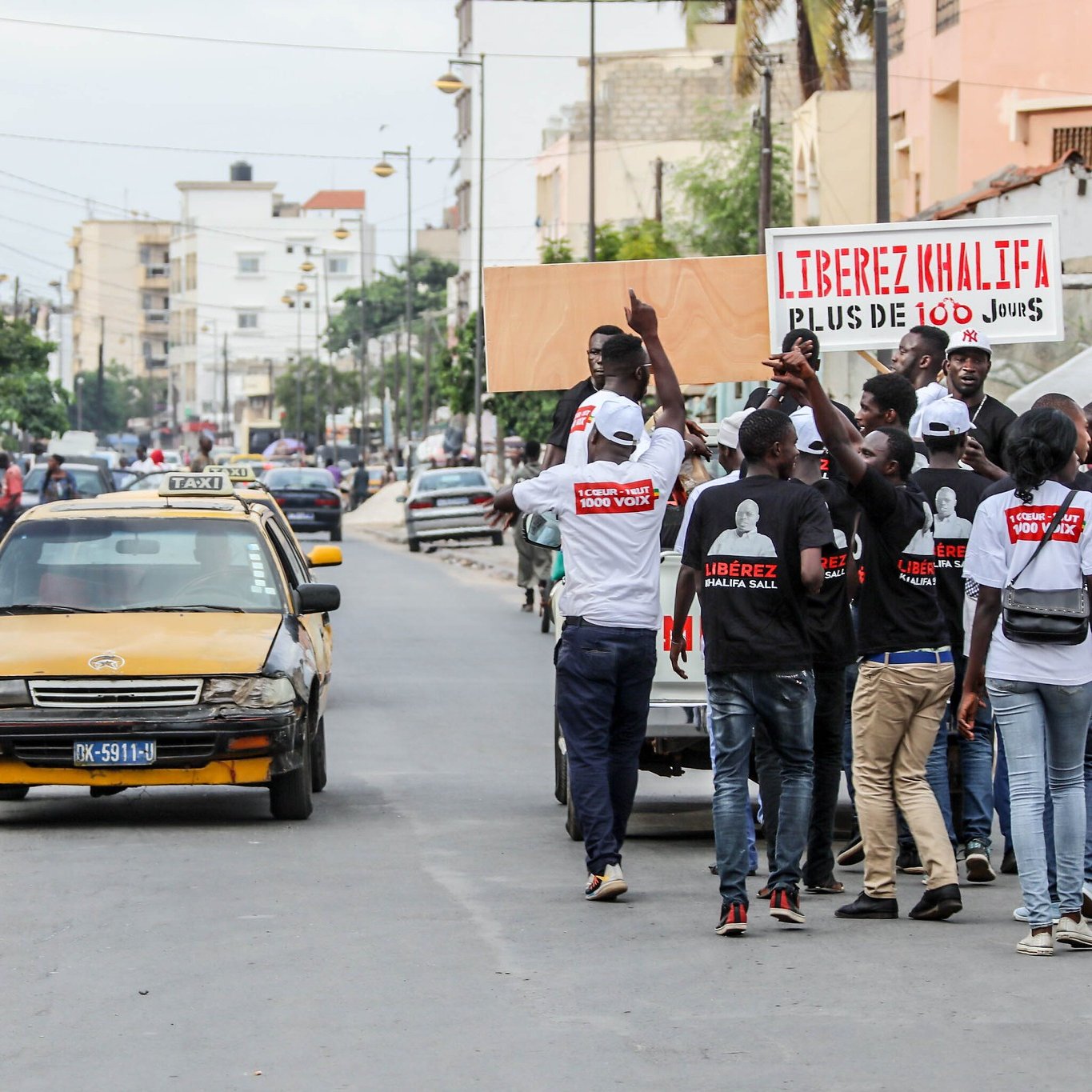 L'image montre une scène animée dans une rue urbaine. À gauche, des personnes marchent, certaines portant des tee-shirts blancs avec des inscriptions visibles. Ils semblent participer à une manifestation ou un rassemblement. Au centre, un taxi jaune est garé sur le côté de la route. Sur le côté droit, il y a plusieurs personnes, et l'ambiance donne une impression de dynamisme et d'engagement. Des affiches sont visibles en arrière-plan, dont l'une mentionne des slogans politiques, suggérant un contexte de revendication ou de mobilisation sociale. Le bruit de la ville, les voix des manifestants et le bruit des véhicules complètent cette atmosphère vivante.