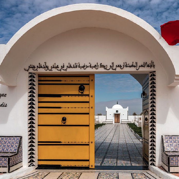 Imaginez un lieu paisible et accueillant, avec une grande porte jaune ornée de motifs géométriques. Cette porte s'ouvre sur un jardin magnifique, symbolisant la beauté et la diversité de la nature. Deux bancs placés de chaque côté de l'entrée, recouverts de carrelage coloré, invitent à la détente. Au-dessus de la porte, une inscription en arabe et en français nous invite à découvrir ce jardin, intitulé "Jardin d'Afrique, Jardin du Paradis". De chaque côté, un drapeau flotte, ajoutant une touche de couleur vive à la scène. Le ciel est lumineux et parsemé de nuages, apportant une lumière douce qui souligne la sérénité de cet endroit. L'ambiance générale est calme et harmonieuse, évoquant un espace de ressourcement et de contemplation.