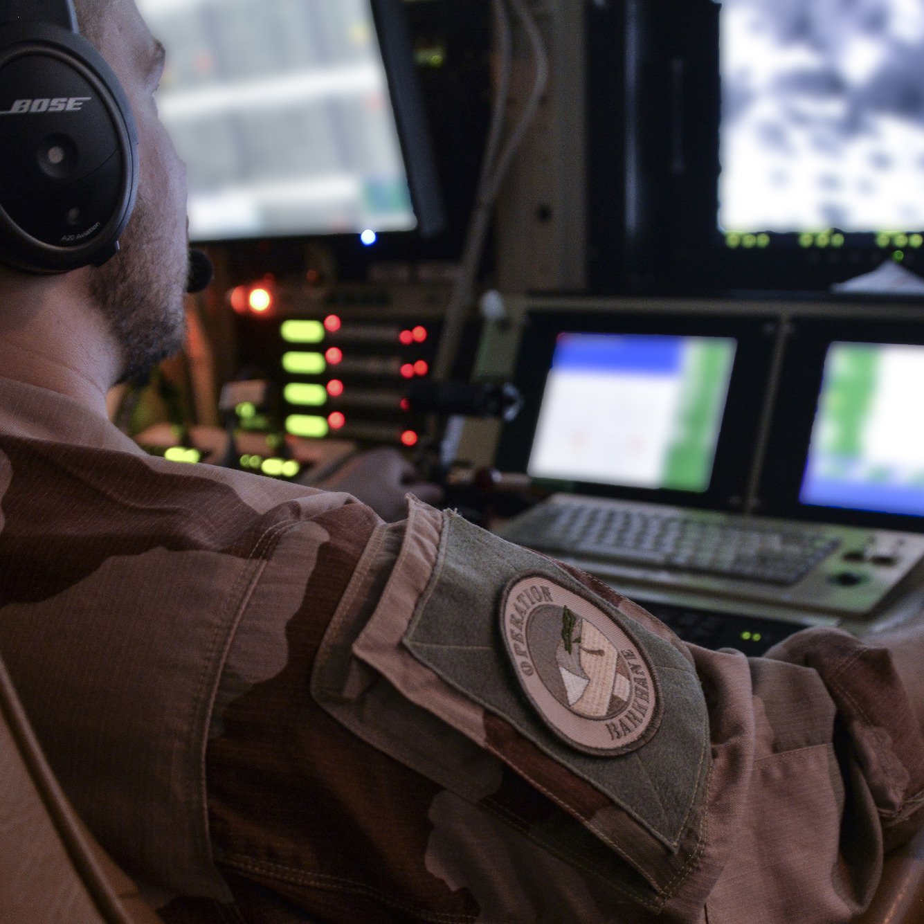 Dans cette image, on voit un homme assis devant un tableau de contrôle, portant un uniforme militaire. Il a un casque sur les oreilles et utilise des manettes et des boutons sur la console. Devant lui, un écran affiche des images en noir et blanc, suggérant qu'il surveille ou contrôle quelque chose à distance. Le décor est sombre, avec des lumières d'indication vertes et rouges autour de lui, ajoutant une atmosphère technologique et militaire à la scène. L'arrière-plan suggère un environnement de travail complexe, probablement dans un avion ou un centre de commandement.