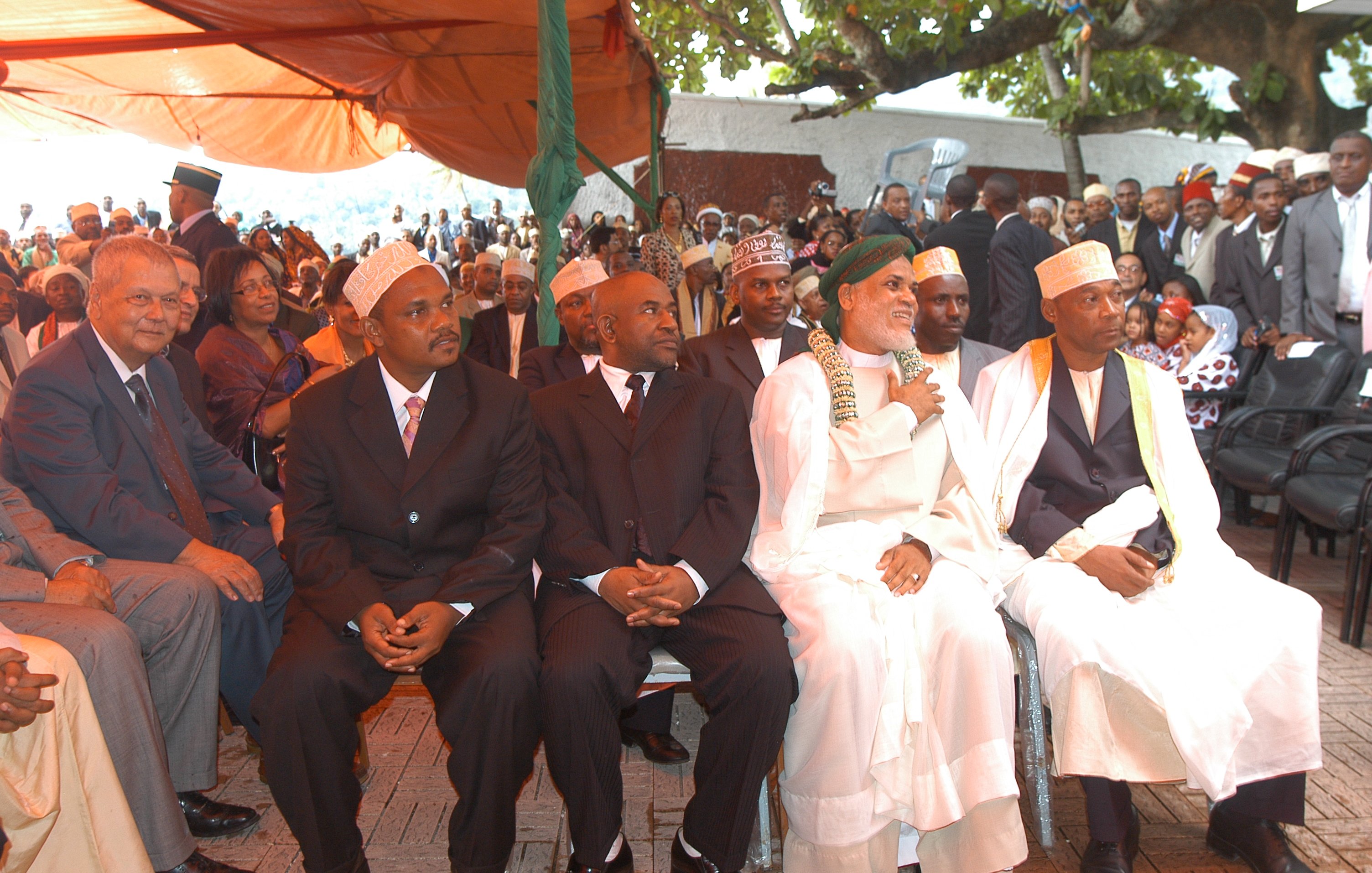 L'image montre un groupe d'hommes assis côte à côte lors d'un événement en plein air. Au premier plan, quatre hommes bien habillés portent des costumes sombres et des chapeaux traditionnels. Le premier homme à gauche a des lunettes et affiche une expression sérieuse. Le deuxième, au milieu, est habillé en noir et semble concentré. Le troisième, portant une robe traditionnelle blanche et un turban, sourit et semble être en conversation. Le quatrième homme, également en tenue traditionnelle, regarde droit devant lui. En arrière-plan, on peut apercevoir une foule de personnes rassemblées, avec des arbres et des tentes fournissant de l'ombre. L'ambiance paraît formelle et respectueuse, suggérant que ce moment est empreint de signification culturelle ou de cérémonie importante.