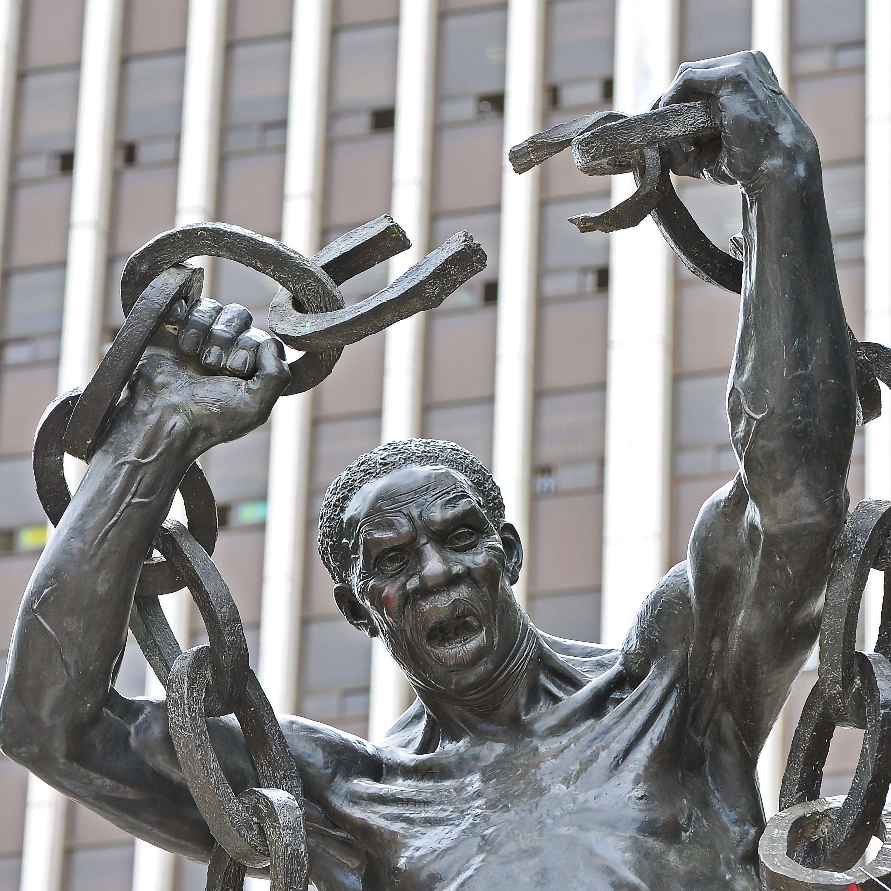 Cette image représente une statue en bronze d'un homme robuste. Il se tient debout, les bras levés au-dessus de sa tête, brandissant de lourdes chaînes brisées, symbolisant la libération et la force. Son visage exprime une intense émotion, avec une bouche ouverte comme s'il poussait un cri de victoire ou de défi. La sculpture est placée devant un bâtiment moderne, avec des lignes verticales qui soulignent contraste entre le passé et le présent. L'œuvre évoque des thèmes de résilience et de lutte pour la liberté.