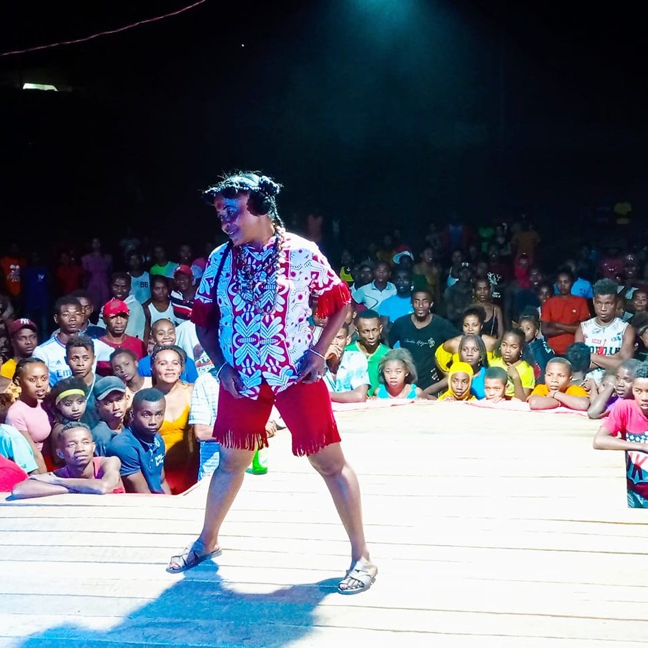 L'image montre une scène vivante et animée. Au premier plan, un artiste, habillé de manière colorée avec une chemise à motifs et un short, se tient sur une scène en bois. Son expression enthousiaste et son attitude dynamique suggèrent qu'il est en plein spectacle. Derrière lui, une foule nombreuse est présente, composée de personnes de tous âges et de toutes origines, certaines souriant et d'autres captivées par la performance. Les lumières autour de la scène créent une atmosphère festive, tandis que l'obscurité en arrière-plan met l'accent sur l'énergie du moment. Les spectateurs semblent engager et réceptifs, témoignant de l'ambiance joyeuse d'un événement en plein air.