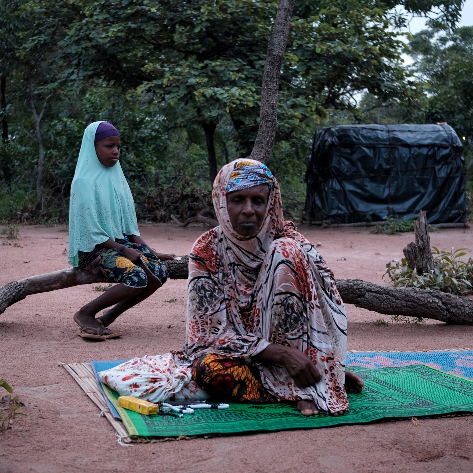 Dans cette image, nous sommes dans un environnement naturel, avec le sol recouvert de terre rouge et entouré d'arbres. À l'avant, une femme est assise sur un tapis, vêtue d'un habit coloré et d'un foulard. Elle a une expression calme. À l'arrière-plan, une jeune fille est assise sur une branche, portant un voile et un vêtement aux motifs colorés. La scène dégage une atmosphère de sérénité et de contemplation, avec la lumière naturelle qui crée une ambiance douce et paisible.