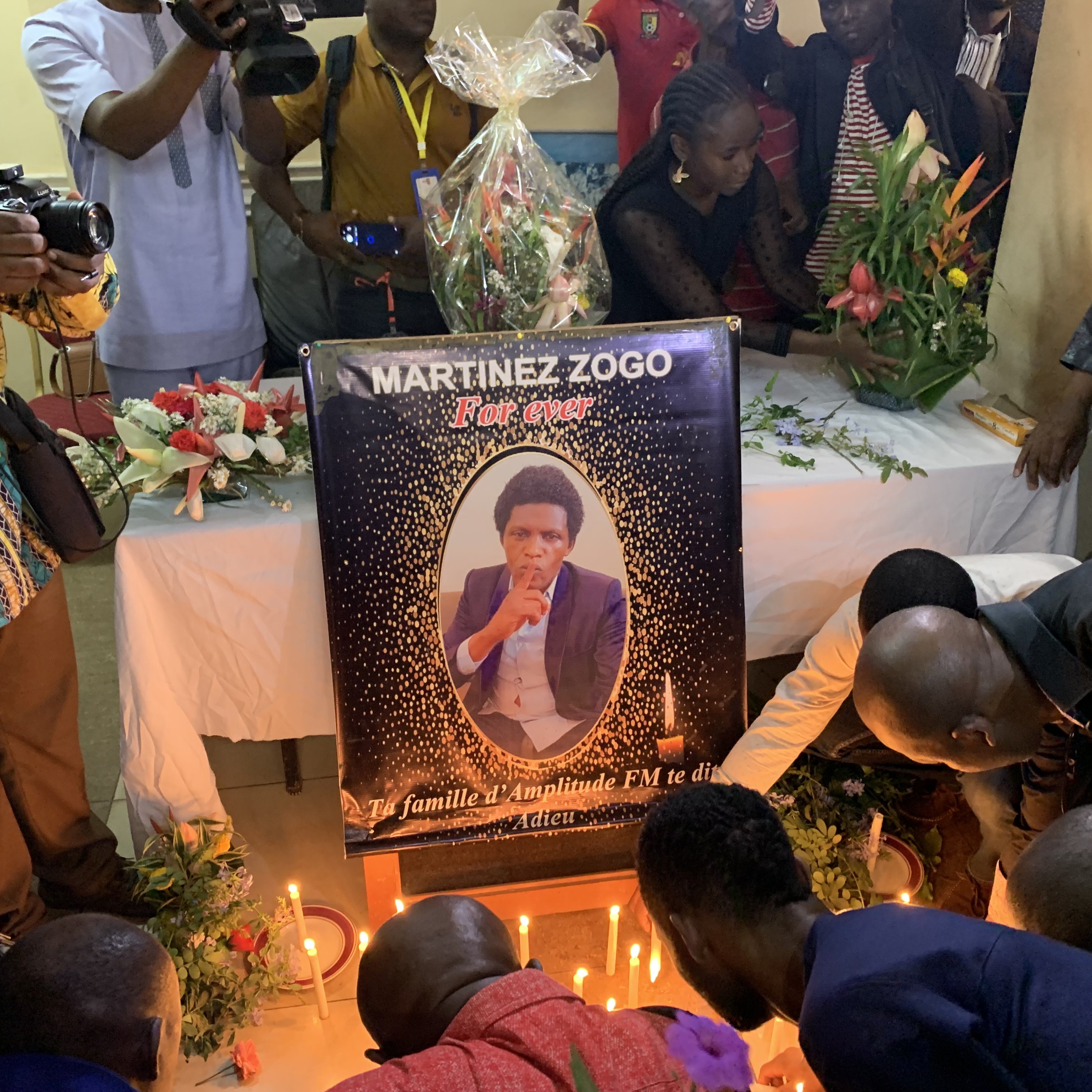 L'image dépeint une scène de recueillement. Au centre, il y a un grand poster représentant Martinez Zogo, encadré de fleurs et de bougies allumées. Le poster montre une photo de lui en train de fumer une cigarette, avec un regard pensif. Autour du poster, plusieurs personnes sont rassemblées, certaines s'agenouillant ou se penchant pour prier. L'atmosphère est solennelle, avec des expressions de tristesse sur les visages. Des fleurs et des bougies décorent la table sur laquelle le poster repose, ajoutant une touche de commémoration à cet hommage.