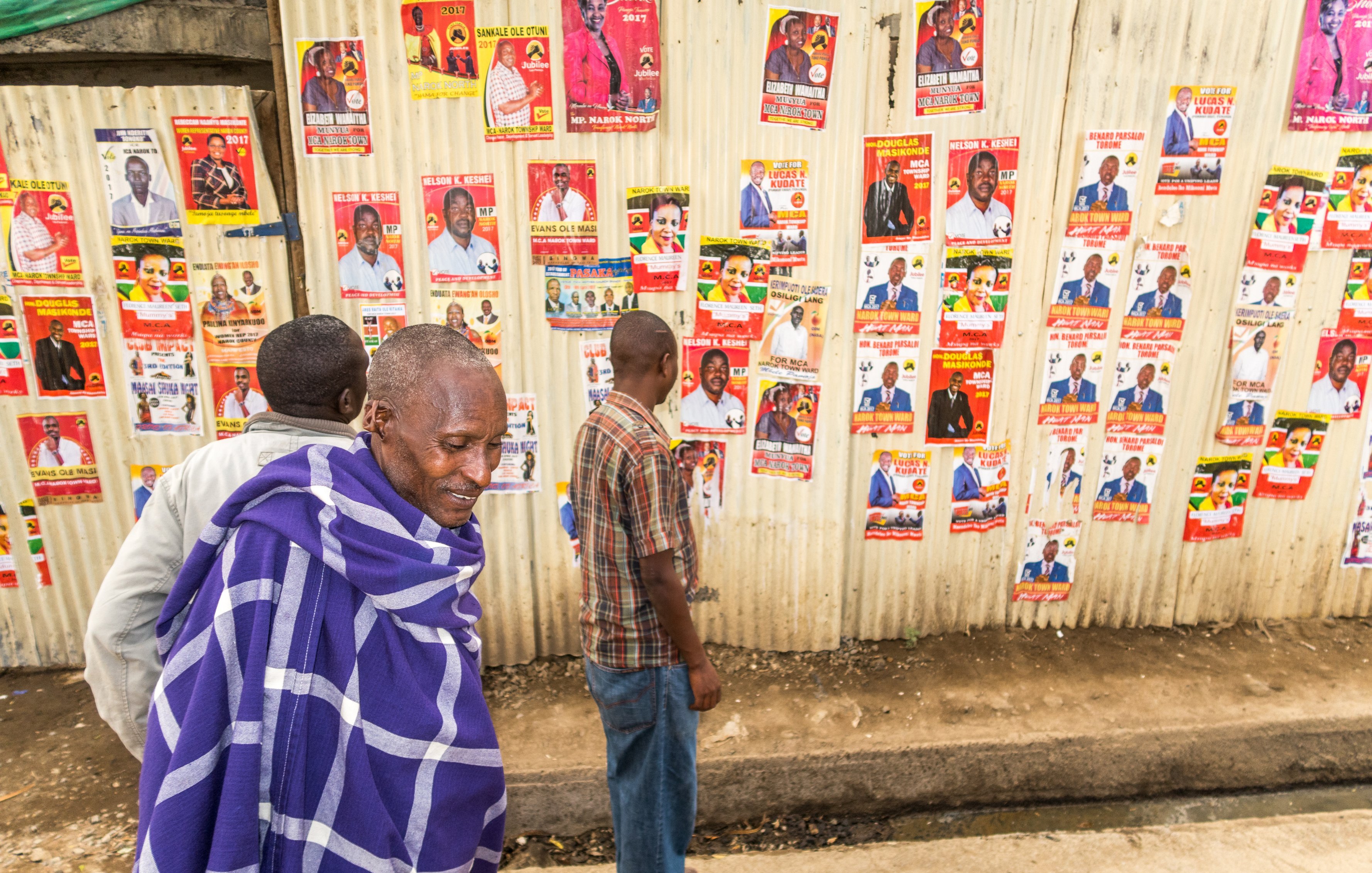 L'image montre un groupe de personnes marchant le long d'un mur recouvert de nombreuses affiches. Ce mur en tôle est tapissé de posters colorés présentant des candidats politiques, avec des visages souriants et des informations écrites. Au premier plan, un homme est vêtu d'un châle traditionnel bleu et blanc, semblant contempler les affiches. À ses côtés, d'autres personnes se déplacent lentement, examinant les posters tandis que le sol est pavé, dans un environnement urbain. L'atmosphère semble à la fois animée et réfléchie, avec un mélange de culture locale et d'engagement politique.