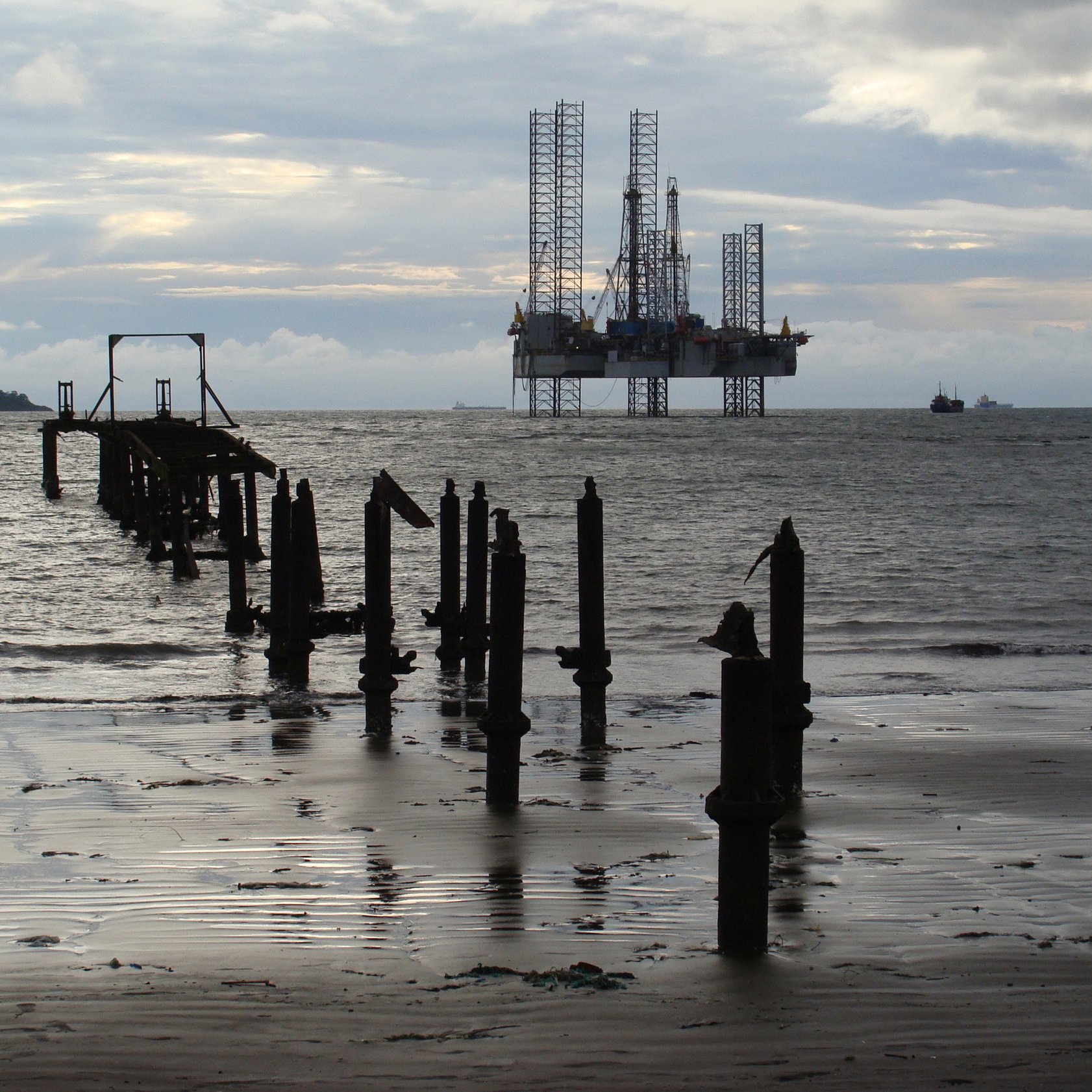 L'image présente un paysage maritime. Au premier plan, on peut voir des restes d'un vieux quai ou d'un embarcadère, avec plusieurs piliers en bois émergent de l'eau. L'eau est calme, et sa surface reflète la lumière, créant une ambiance paisible. À l'arrière-plan, une plateforme pétrolière se dresse majestueusement, avec ses grandes structures métalliques et ses grues, accentuant le contraste entre la nature et l'infrastructure industrielle. Le ciel est nuageux, laissant entrevoir une palette de gris, avec des nuances plus claires et plus sombres. L'ensemble de la scène évoque un mélange de sérénité et de robustesse, témoignant de l'interaction entre l'homme et l'environnement marine.