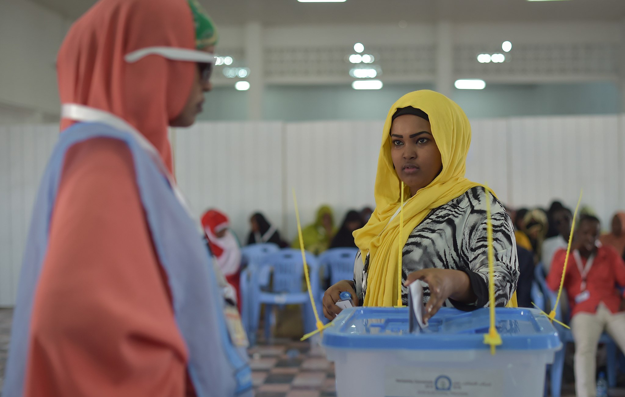 L'image montre une scène de vote dans un lieu organisé. À gauche, une femme porte un hijab orange et des lunettes de soleil, regardant vers une autre femme, qui est située à droite. Cette dernière, en hijab jaune, semble concentrée alors qu'elle dépose son bulletin dans une urne transparente devant elle. L'intérieur de la salle est éclairé, avec des chaises en plastique bleues disposées en rangées. On peut percevoir une atmosphère de participation et d'engagement civique. Des femmes en arrière-plan sont également présentes, créant une ambiance communautaire.