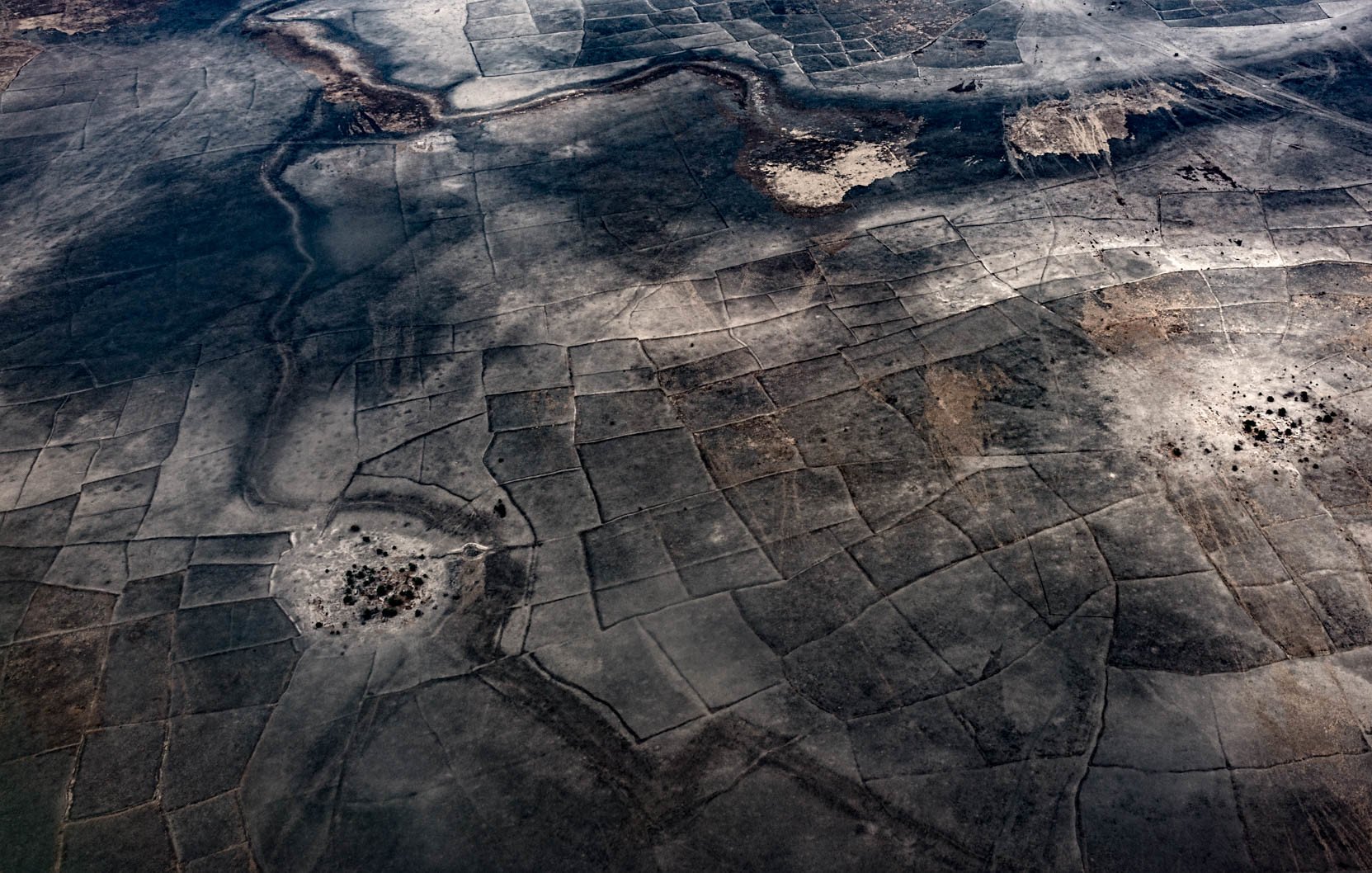 L'image montre un paysage vaste et aride vu d'en haut, avec des motifs géométriques formés par des parcelles de terre. Les teintes dominantes sont des nuances de gris et de marron, évoquant un terrain desséché, peut-être dans une région désertique. On y perçoit des zones plus sombres, qui pourraient représenter des végétations ou des matériaux différents. Les formes géométriques créent une texture visuelle intéressante, tandis que le sol semble craquelé par un manque d'eau. Cette scène dégage une sensation de vastitude et de tranquillité, tout en reflétant une nature rude et indomptée.