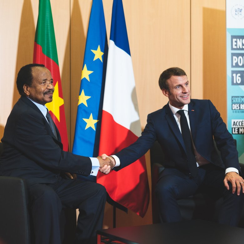 Dans cette image, deux hommes sont assis l'un en face de l'autre, se serrant la main dans un geste formel. L'homme à gauche porte un costume sombre et a une expression souriante. À sa droite, un autre homme, également en costume, affiche un sourire poli. En arrière-plan, on peut noter deux drapeaux : l'un est le drapeau du Cameroun, l'autre le drapeau français et le drapeau de l'Union européenne. L'environnement semble être un espace de réunion officiel, avec des panneaux informatifs visibles. L'atmosphère dégage une impression de cordialité et de coopération.