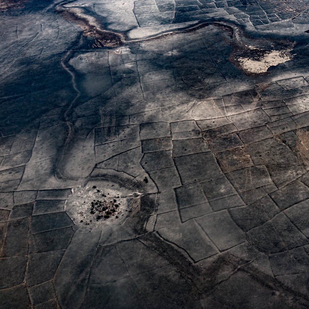 L'image montre un paysage vaste et aride vu d'en haut, avec des motifs géométriques formés par des parcelles de terre. Les teintes dominantes sont des nuances de gris et de marron, évoquant un terrain desséché, peut-être dans une région désertique. On y perçoit des zones plus sombres, qui pourraient représenter des végétations ou des matériaux différents. Les formes géométriques créent une texture visuelle intéressante, tandis que le sol semble craquelé par un manque d'eau. Cette scène dégage une sensation de vastitude et de tranquillité, tout en reflétant une nature rude et indomptée.
