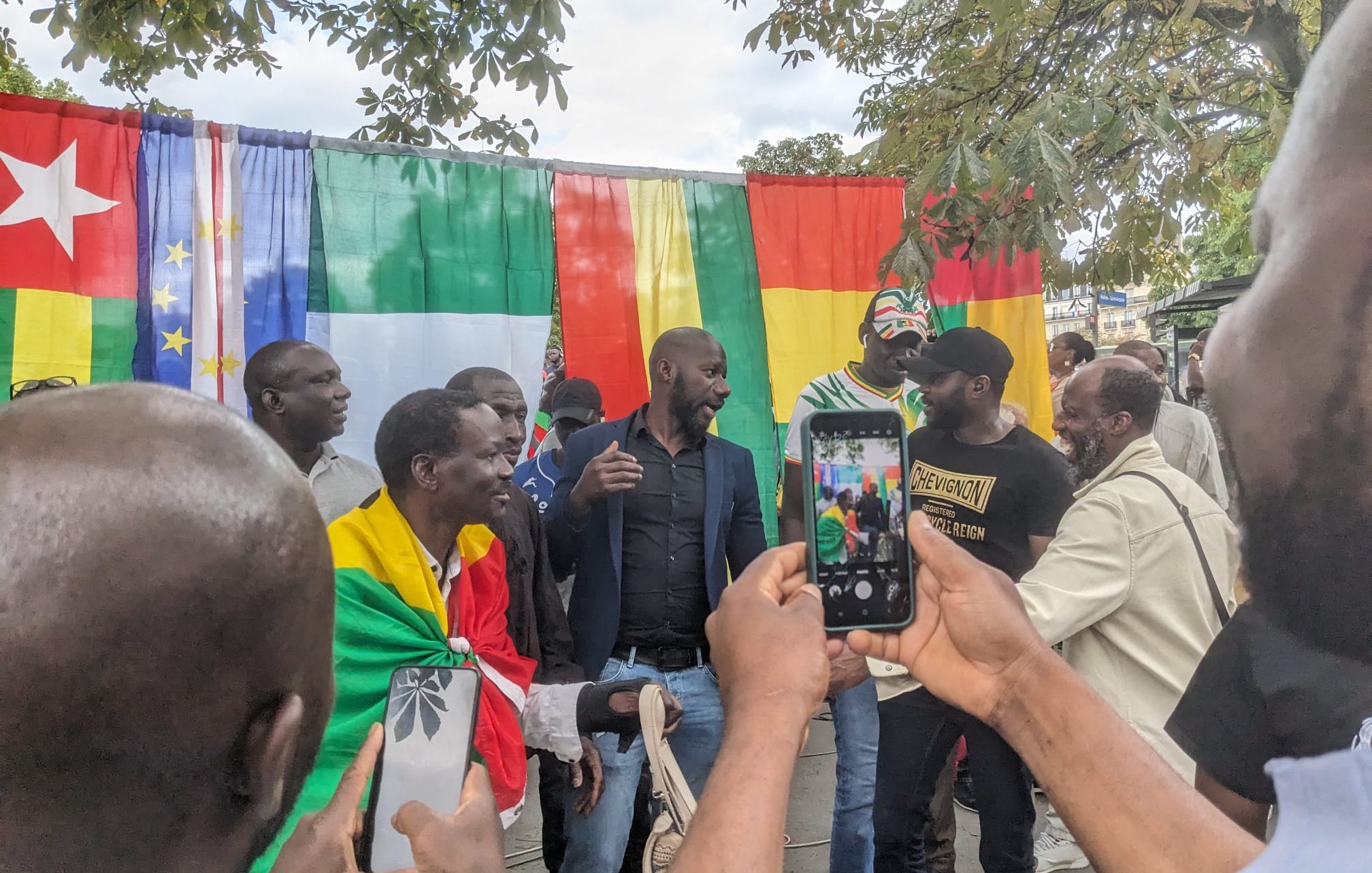 L'image montre un groupe de personnes réunies autour d'un fond coloré constitué de drapeaux. Les drapeaux affichés incluent ceux du Cameroun, de l'Union européenne, du Mali et de la République de Guinée. Des hommes portent des vêtements variés, certains drapés dans des drapeaux, ce qui suggère un événement festif ou culturel. Ils semblent en discussion animée, souriant et échangeant des plaisanteries. Au premier plan, un participant tient un téléphone portable, probablement pour capturer le moment. L'atmosphère semble conviviale et pleine d'énergie.